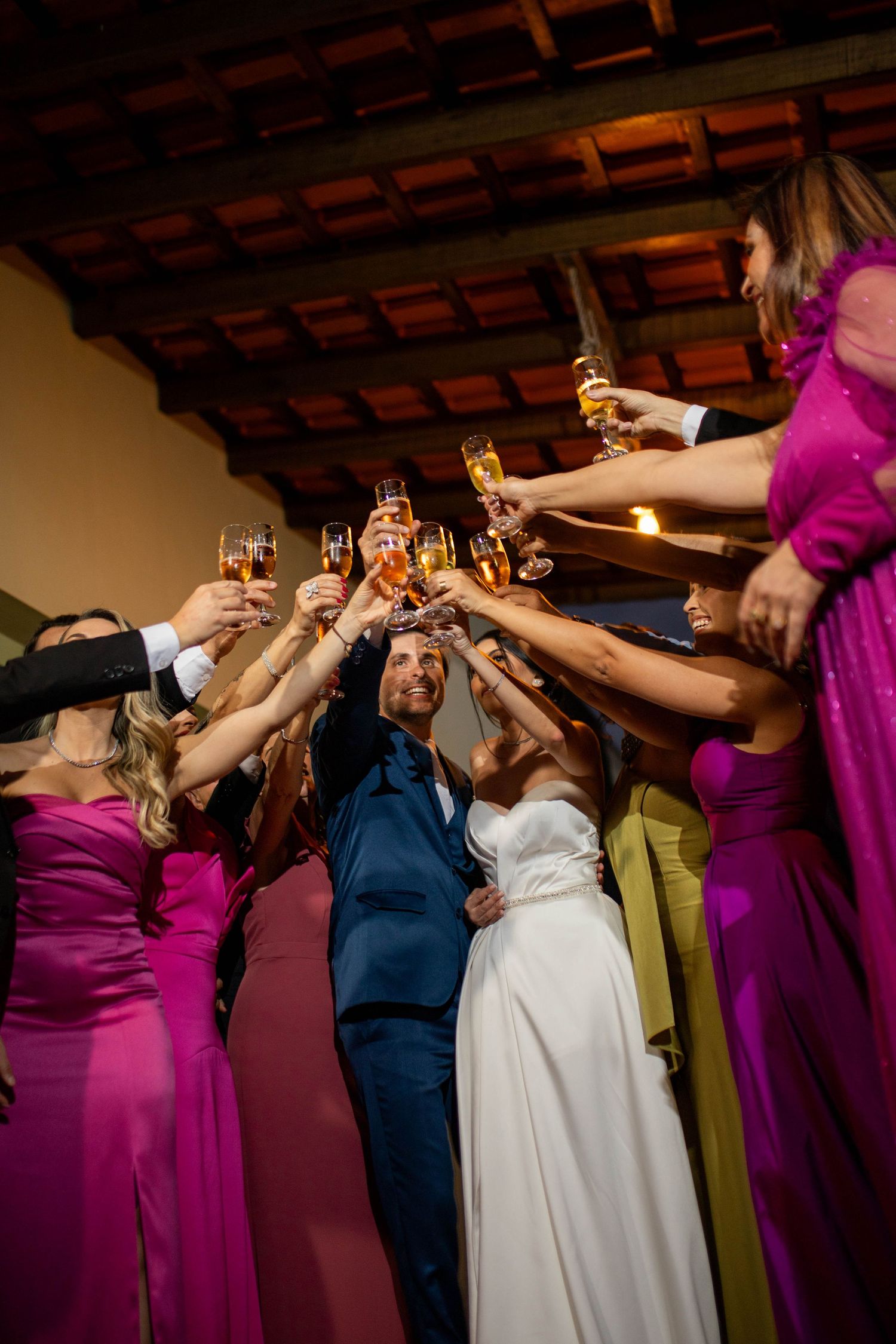 Bride and groom toasting champagne with wedding guests in pink gowns at elegant indoor reception celebration.