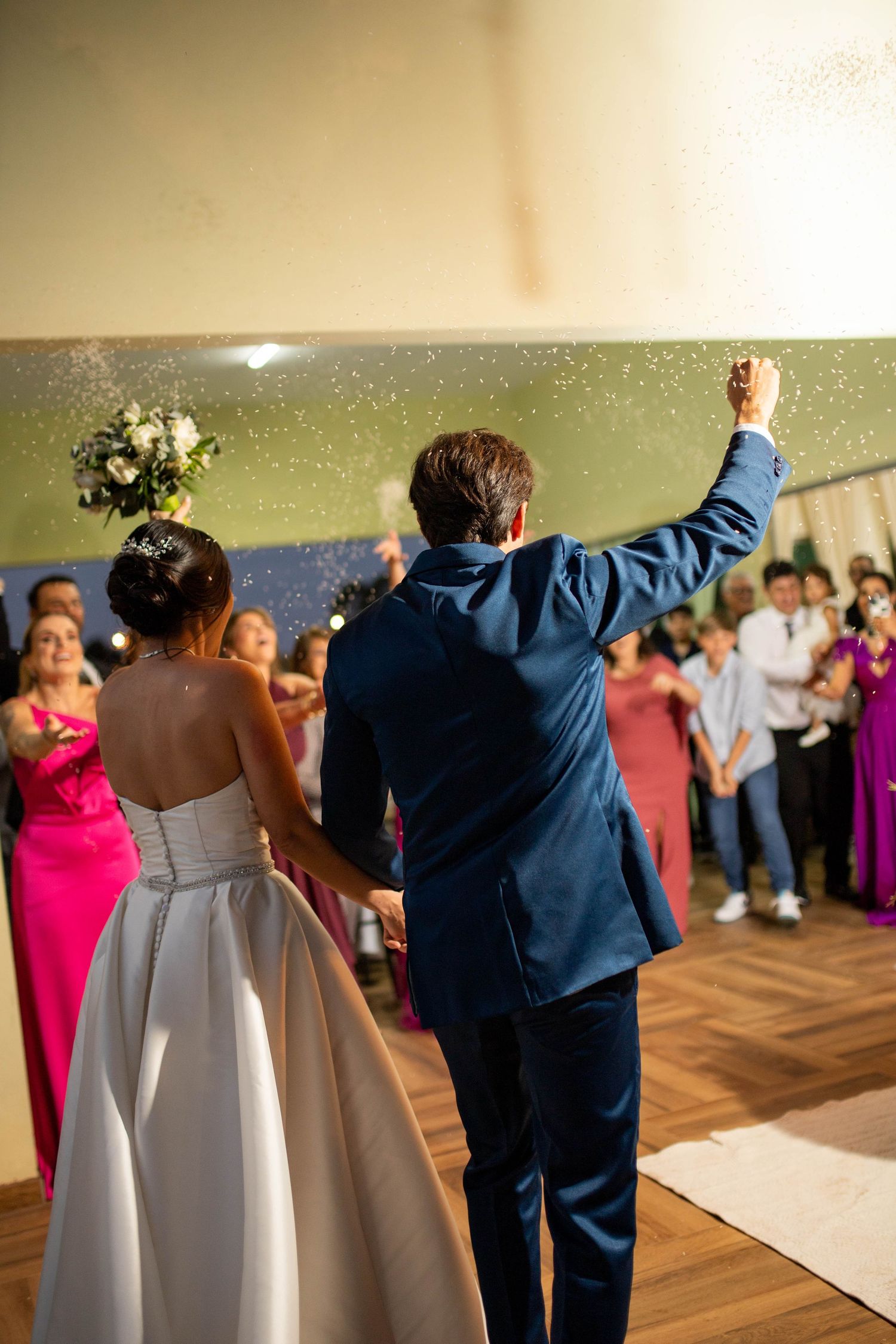 Bride and groom entering wedding reception, holding hands with arm raised in celebration, guests applauding in background.