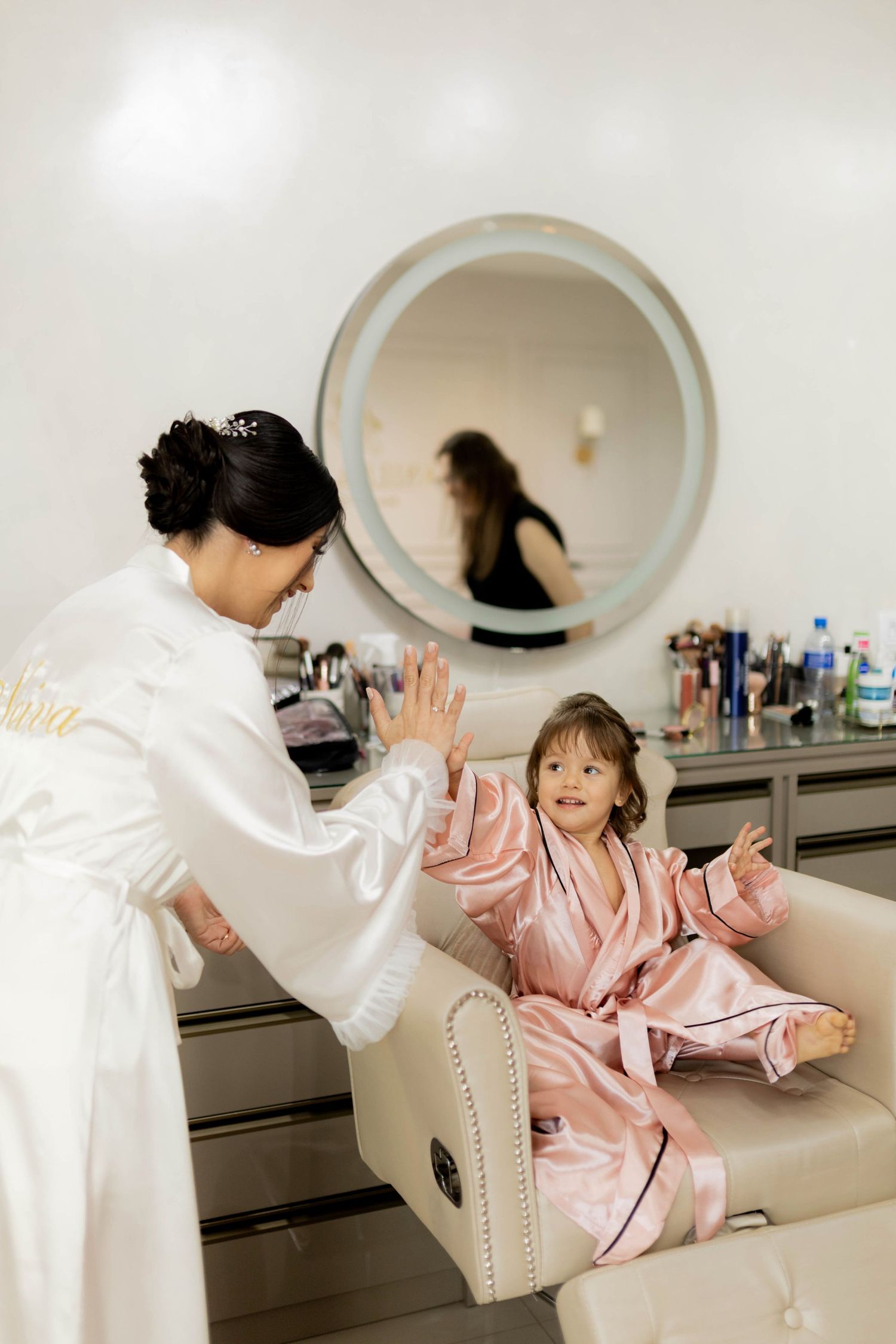 Bride in white robe plays with young girl in pink satin robe at bridal suite vanity mirror.