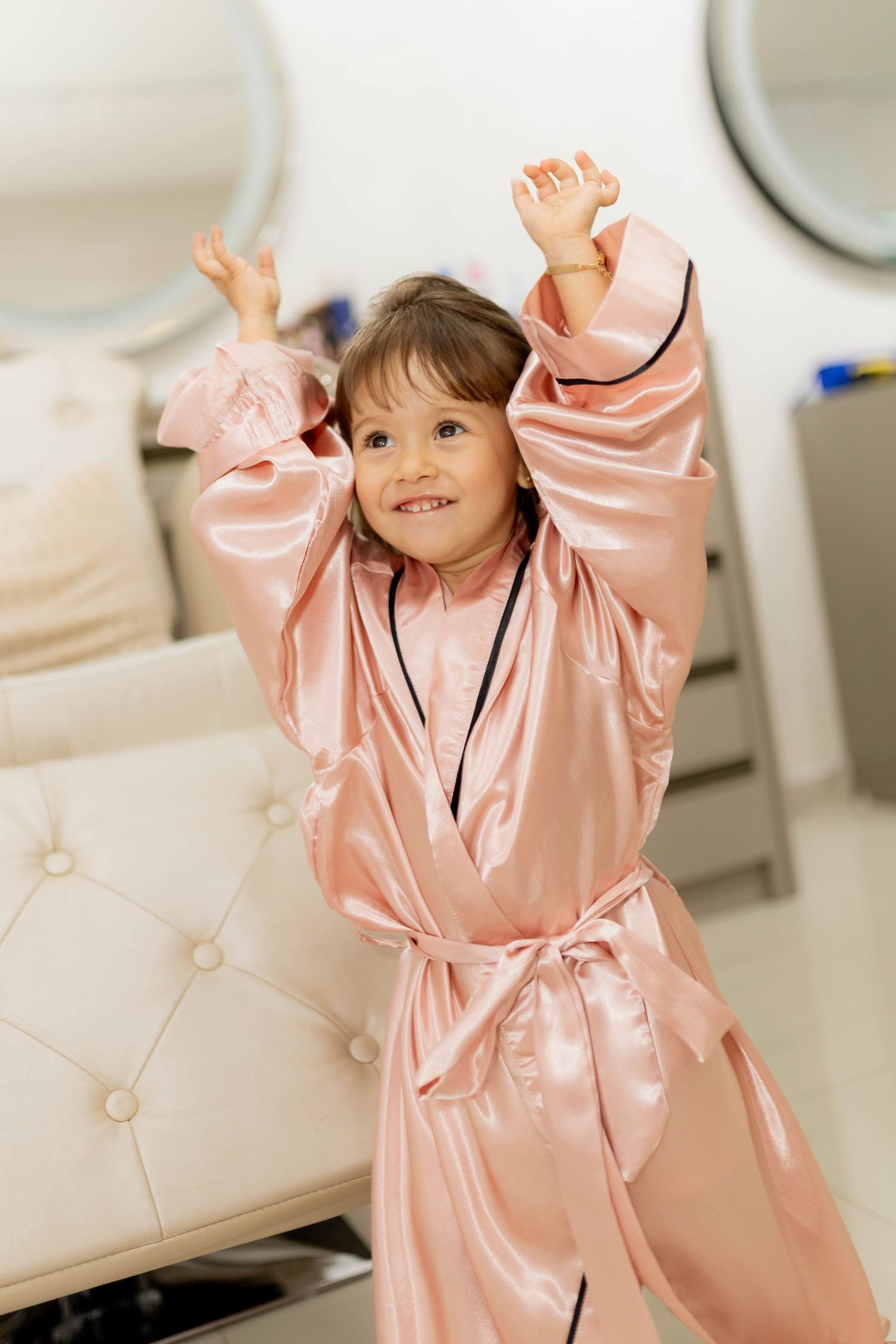 Young girl in oversized pink satin robe smiling with arms raised playfully in a bright bedroom.