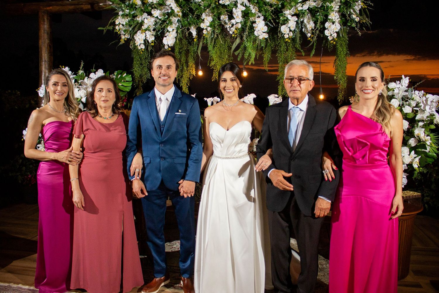 Wedding party poses together, bride in white gown, groom in blue suit, flanked by family in pink formal attire.