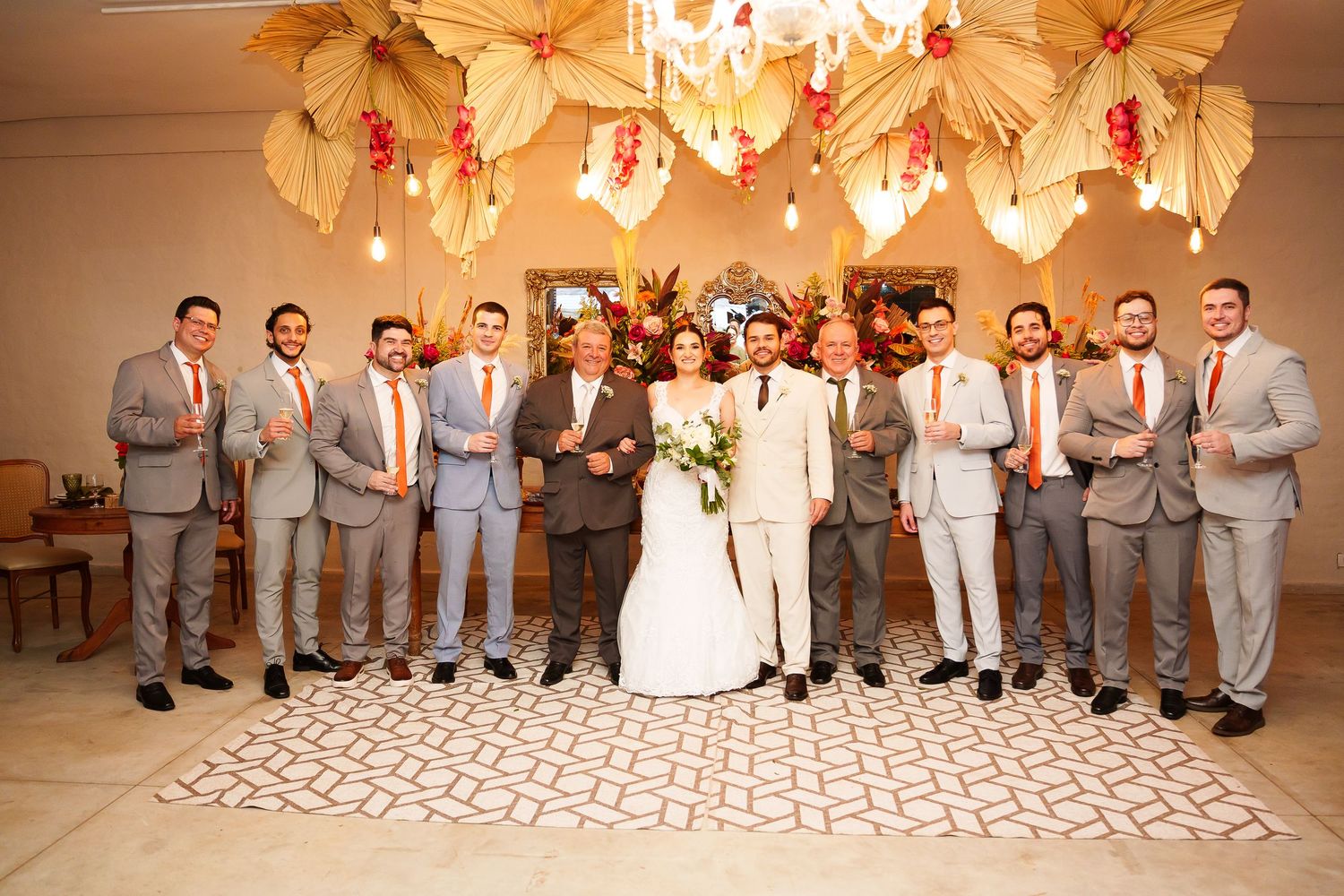 Wedding party posing indoors with bride and groom, groomsmen in gray suits with orange ties, floral ceiling decor.
