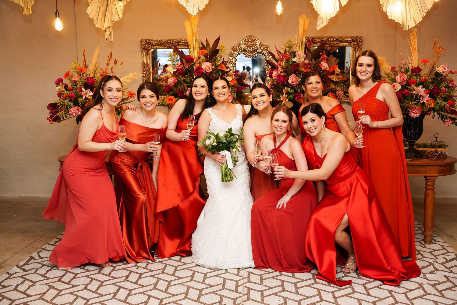 Bride in white gown poses with seven bridesmaids in red satin dresses, surrounded by vibrant floral arrangements.