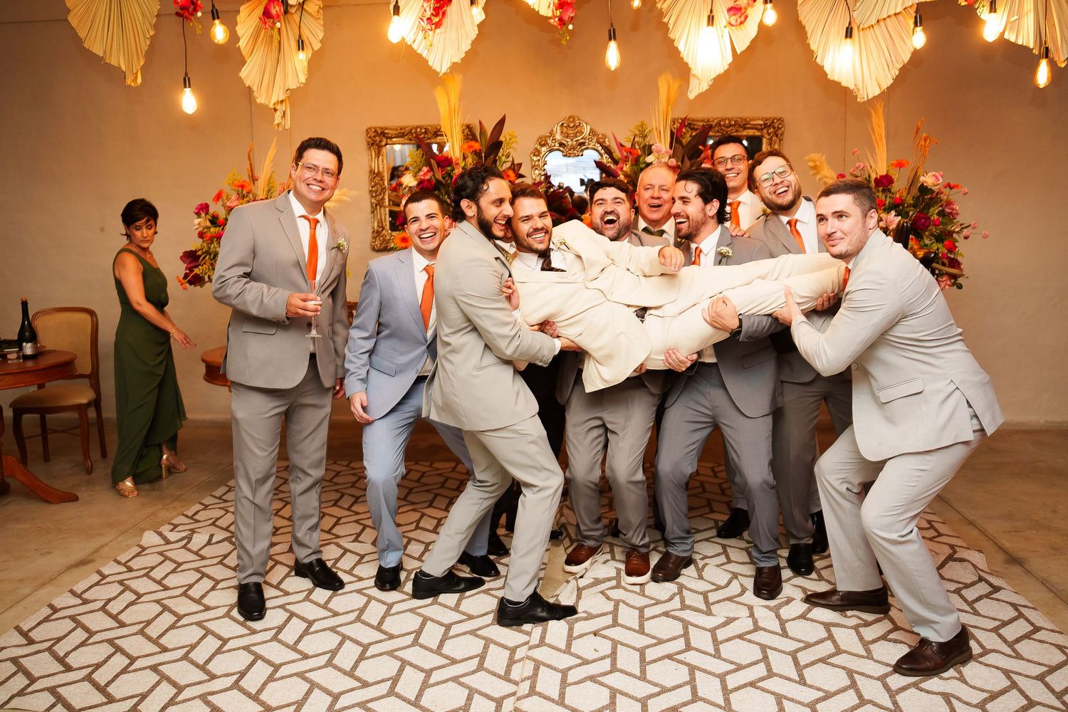 Groomsmen in gray suits lifting the groom at a wedding reception with fall floral decor.