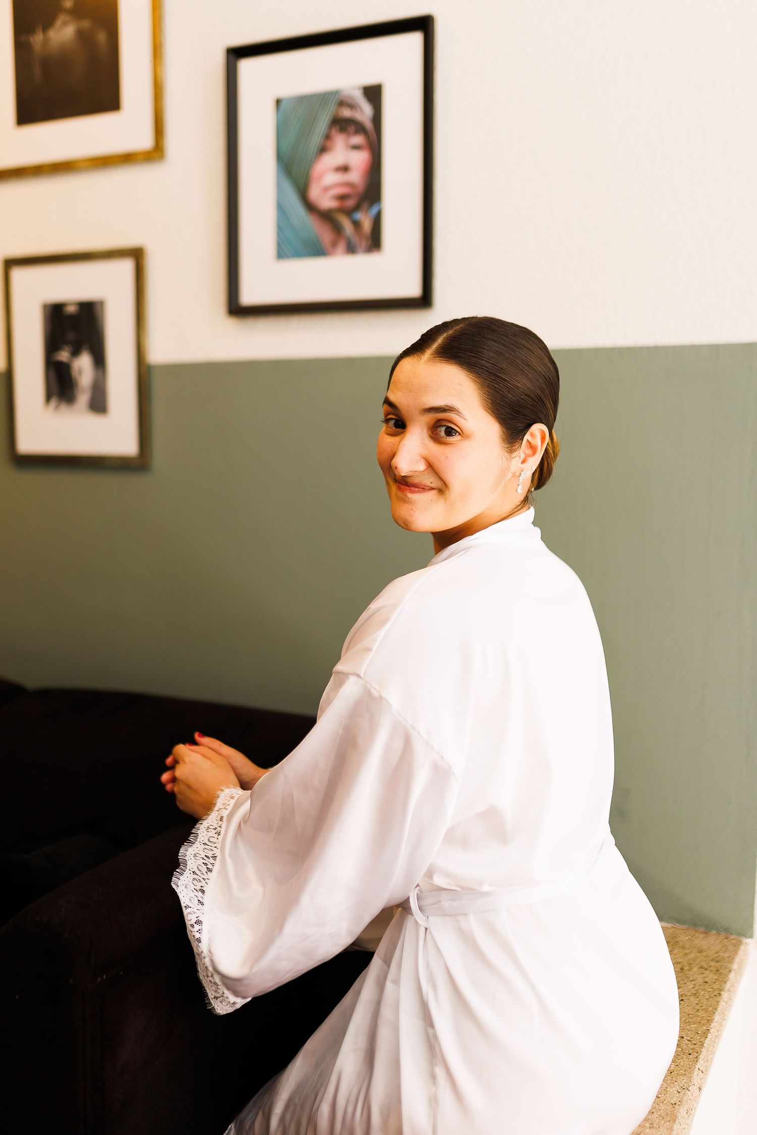 Woman in white lace robe sitting and smiling over shoulder, framed portraits on sage green wall behind her.