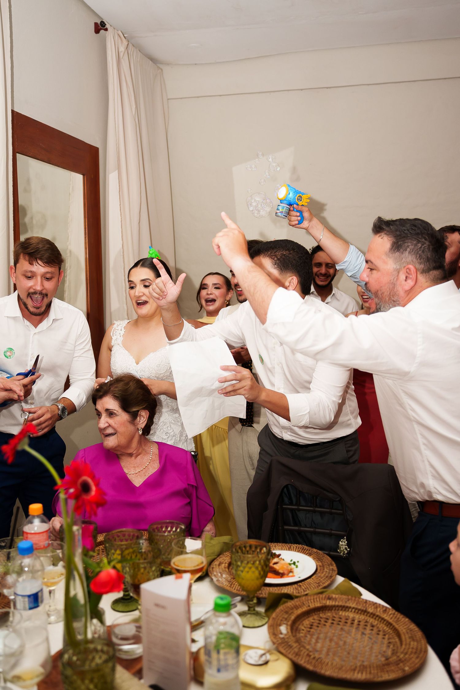 Joyful wedding reception with guests celebrating, bride in white dress, people holding water guns, festive table setting.