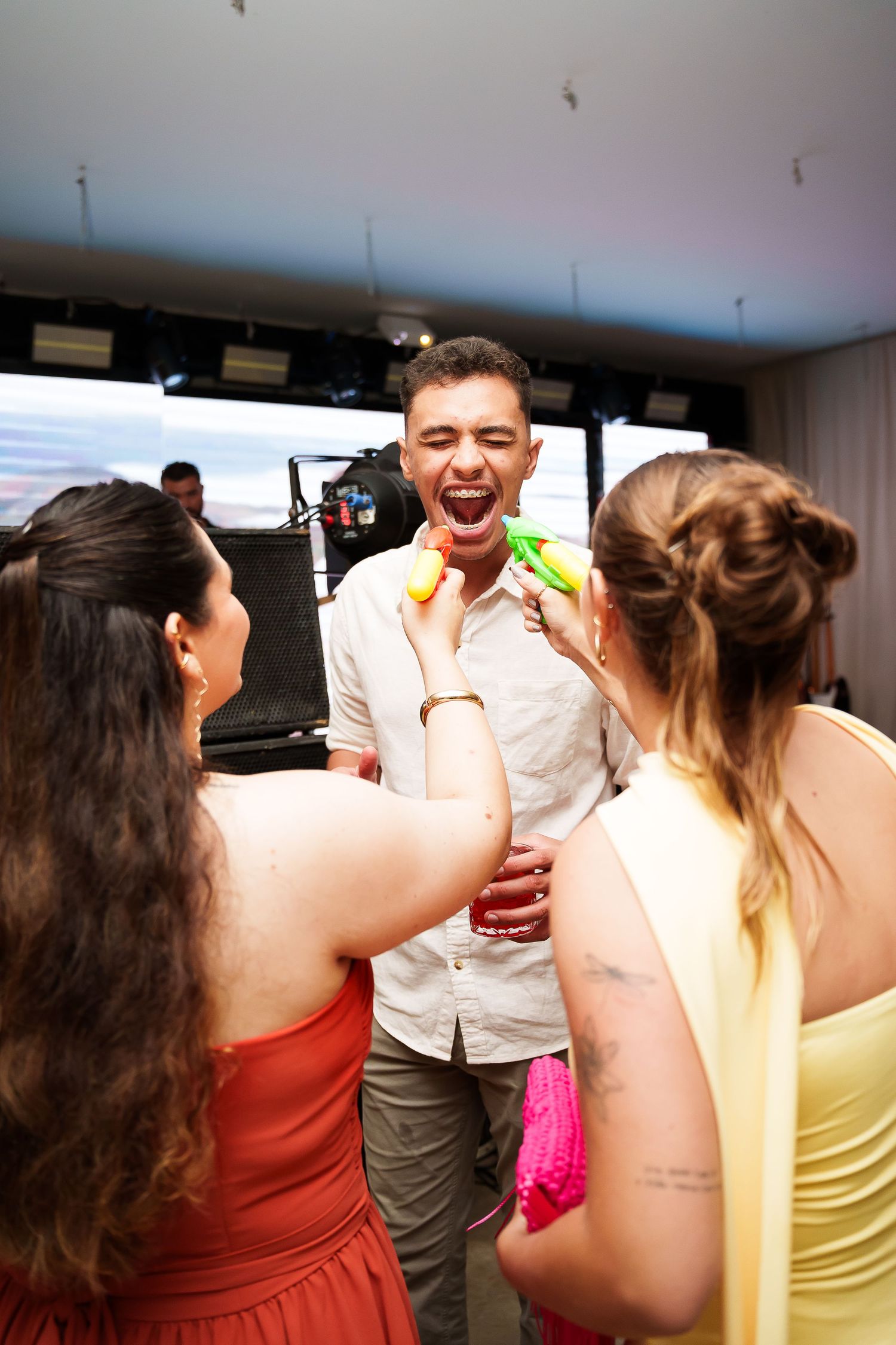 Two women playfully shooting water guns at a laughing man at a lively indoor party celebration.
