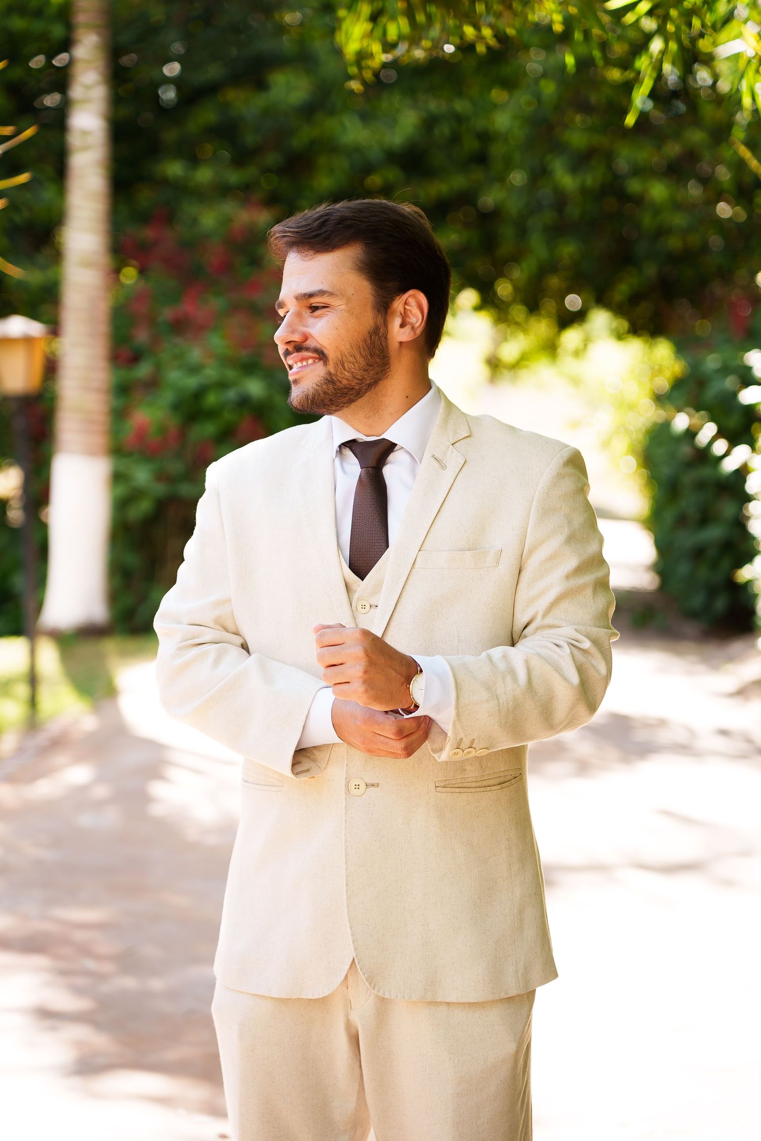 Smiling groom in beige three-piece suit adjusting cufflinks outdoors with lush green trees in background.