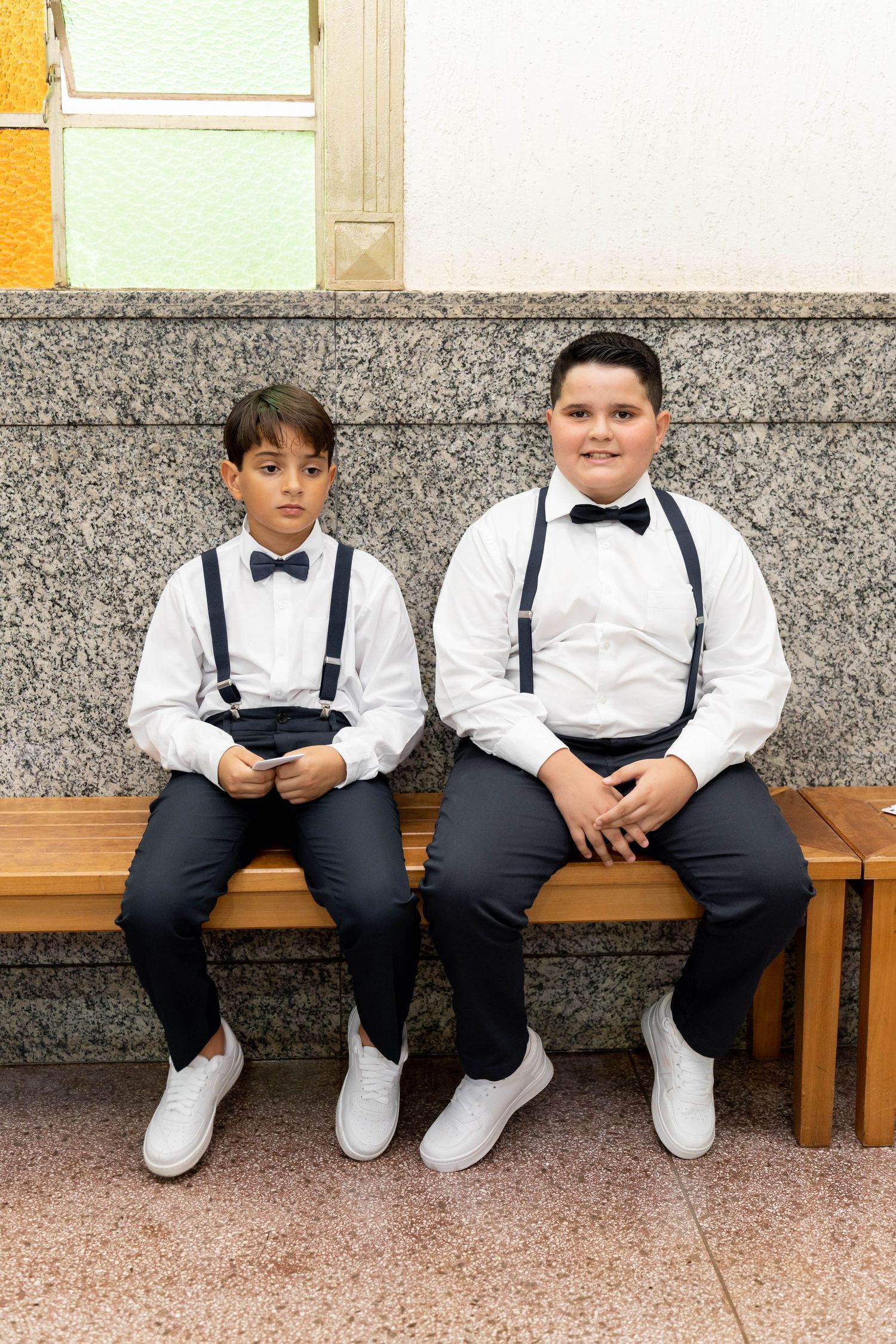 Two boys in matching white shirts, bow ties, suspenders, and black pants sit on a wooden bench.