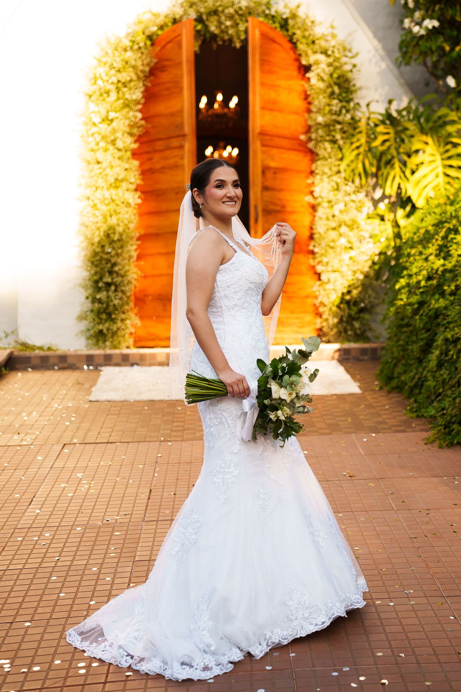 Elegant bride in white lace mermaid gown holding bouquet, standing before floral-adorned wooden doors.