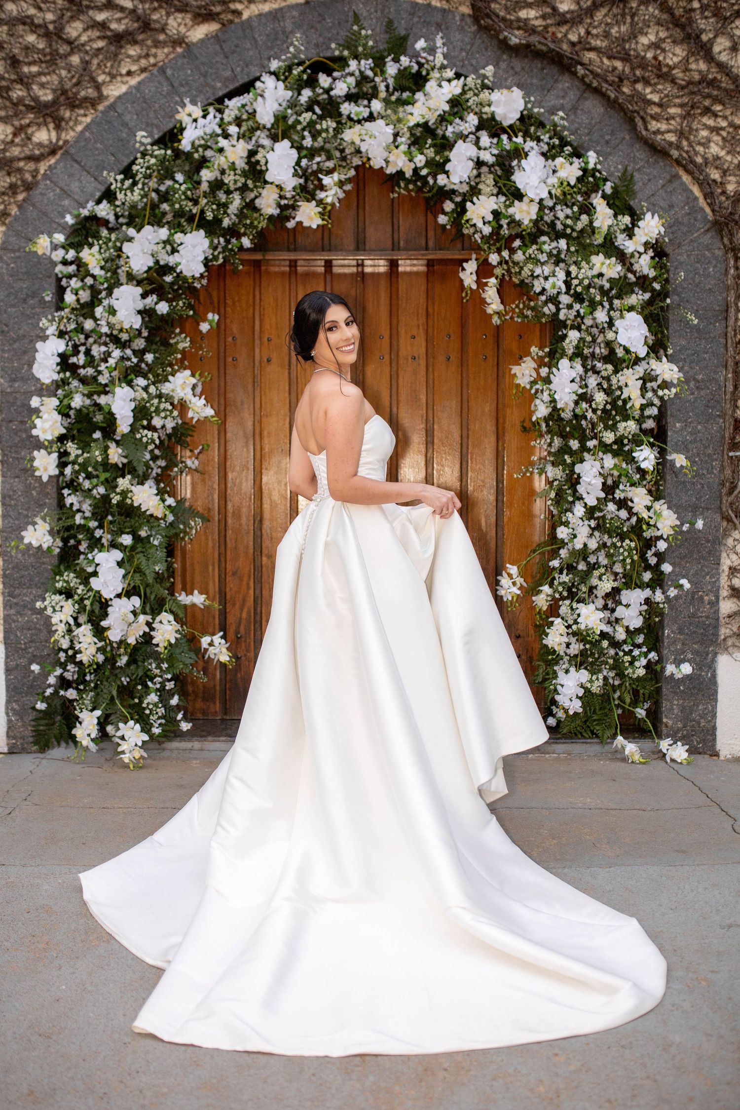 Bride in elegant white satin ball gown poses under floral arch with white roses at wooden church door.