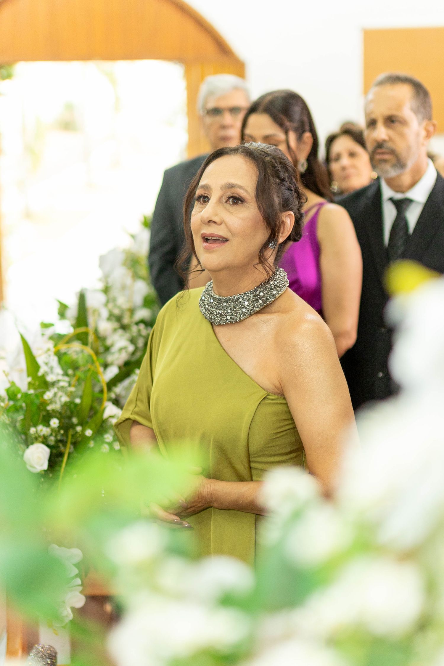 Woman in olive green gown with crystal necklace at a wedding ceremony surrounded by white floral arrangements.