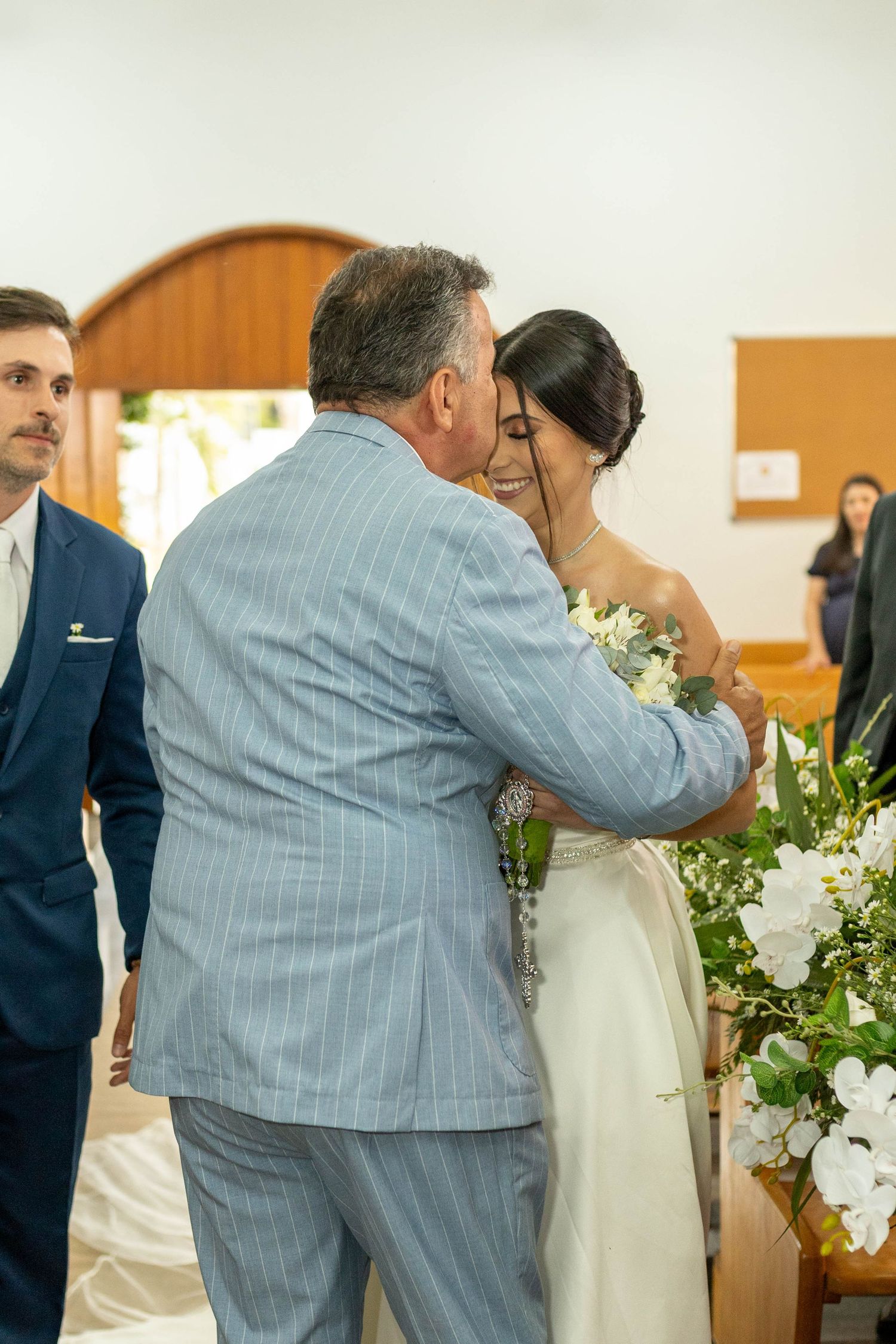 Father of the bride sharing an emotional hug with his daughter in a white wedding gown inside a church.