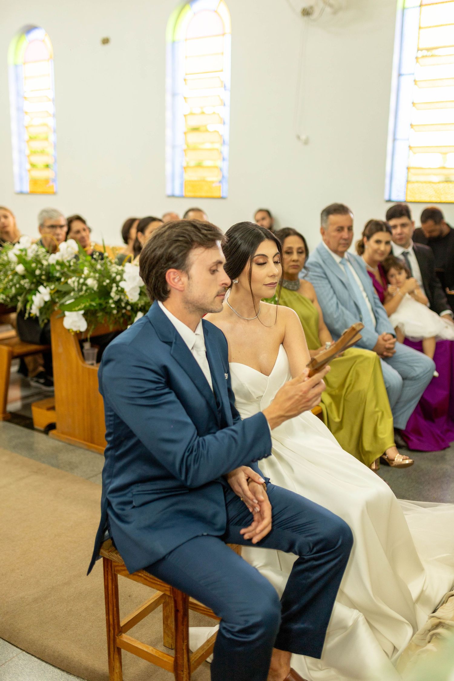 Bride and groom sitting at church altar during wedding ceremony, guests seated in pews behind them.