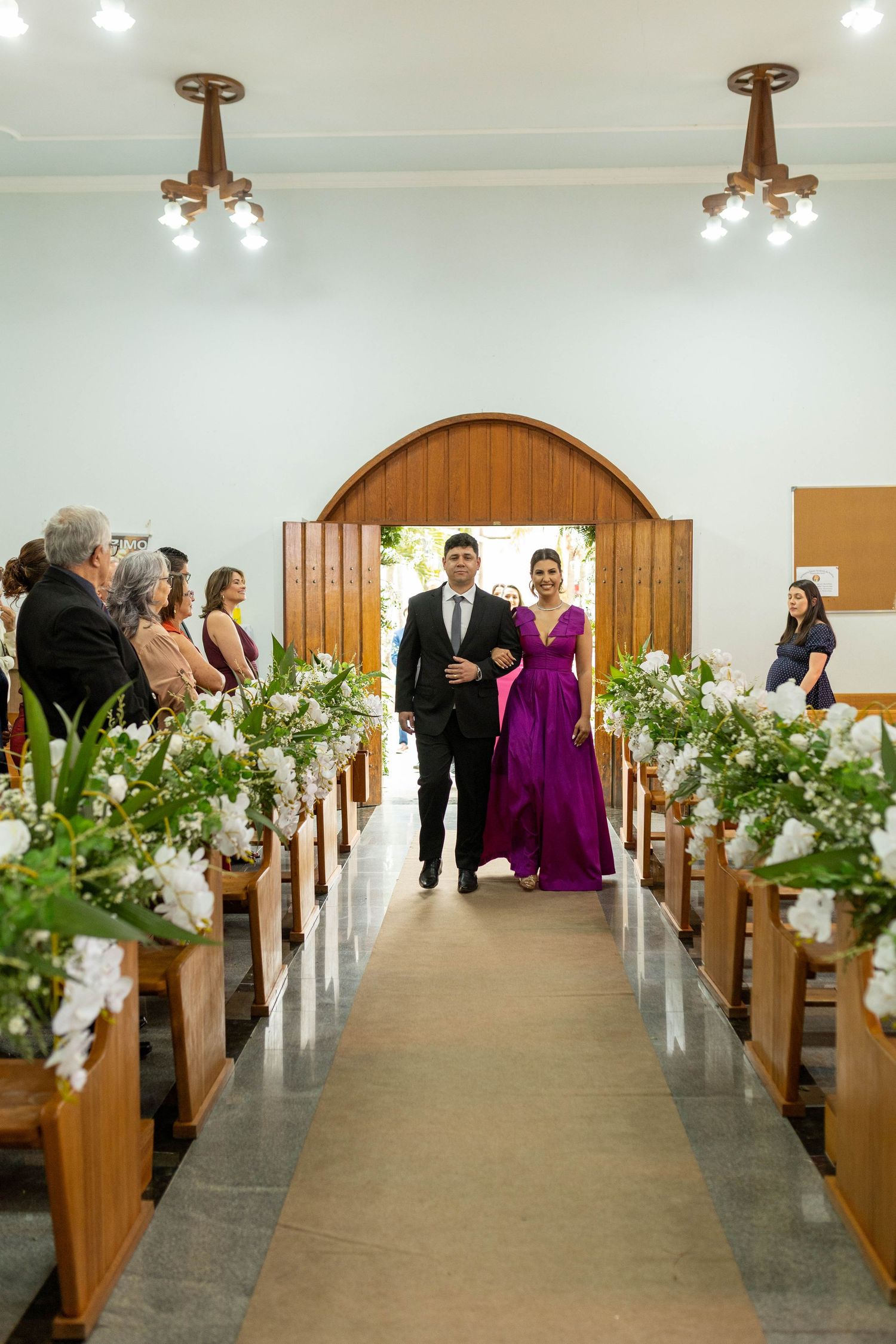 A couple walks down a flower-adorned church aisle, the woman in a purple gown, during an elegant wedding ceremony.