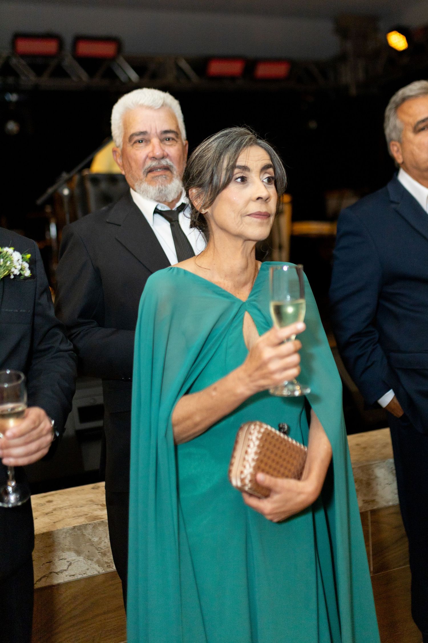 Elegant woman in teal cape gown holding champagne glass at formal event with guests in background.