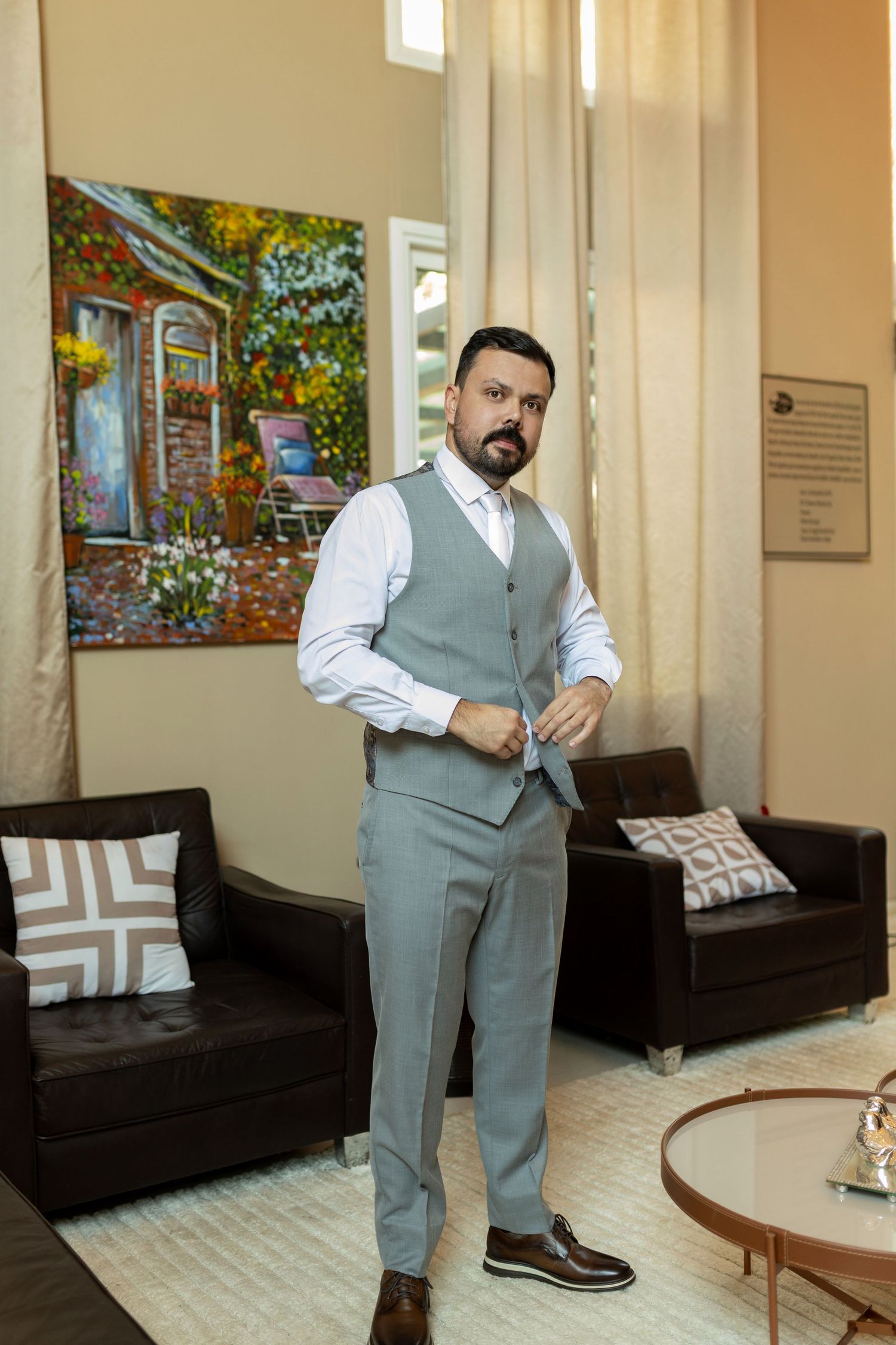 Man in gray vest suit adjusting buttons in elegant room with colorful painting and leather sofas.
