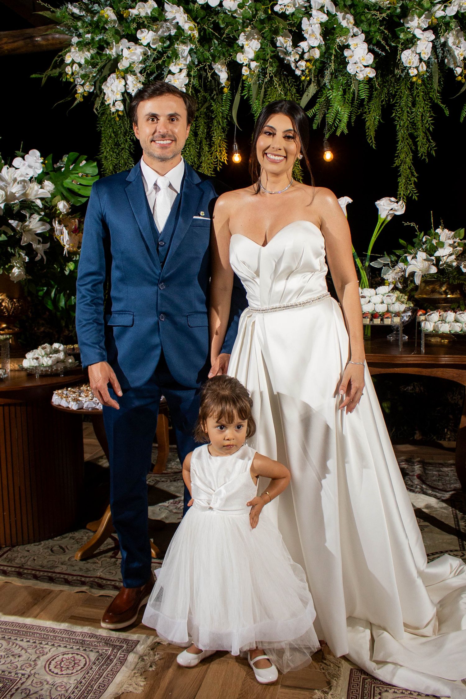 Bride in white gown and groom in blue suit pose with flower girl at elegant floral wedding ceremony.