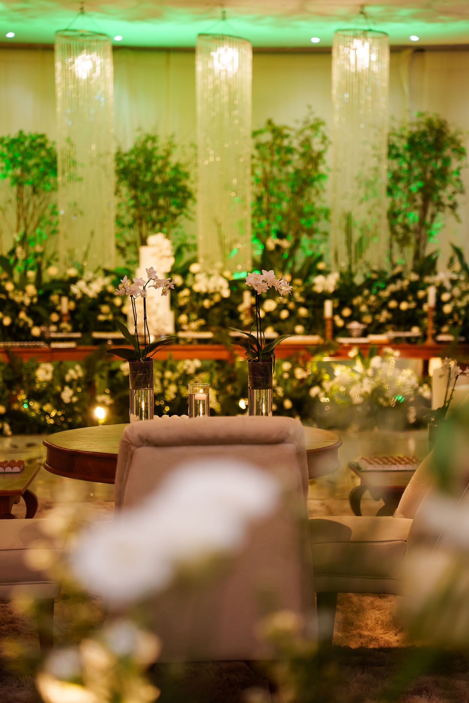 Elegant wedding reception venue with orchids, white flowers, green trees, and sparkling chandelier curtains in warm lighting.