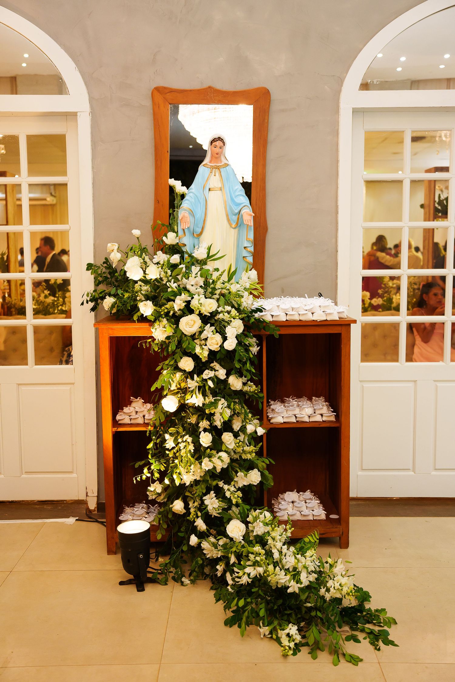 Elegant floral arrangement with white roses cascading down a wooden shelf beside a Virgin Mary statue and mirror.