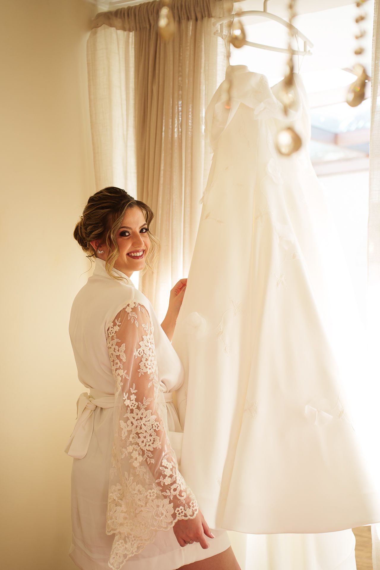 Smiling bride in lace robe standing next to her white wedding dress hanging by a sunlit window.