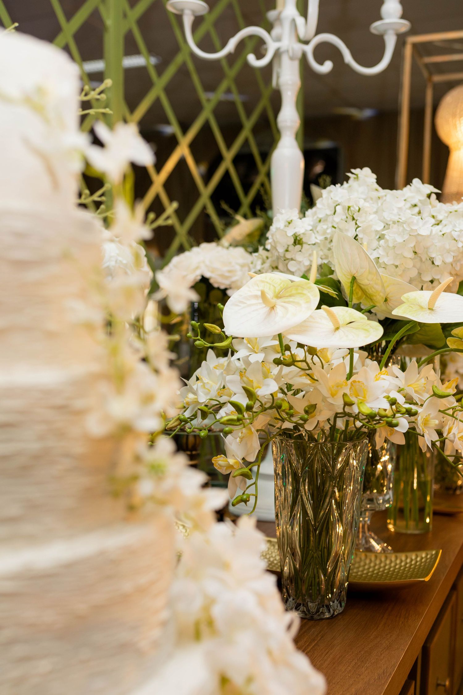 Elegant wedding cake beside white floral arrangements with hydrangeas and anthuriums in crystal vases on a wooden table.