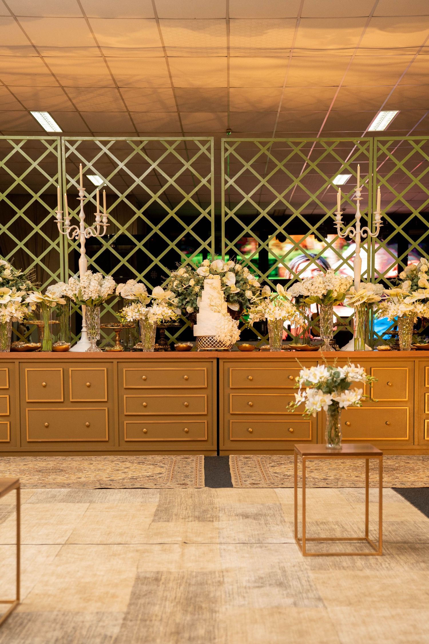 Elegant wedding dessert table with white floral arrangements and tiered cake on gold dressers with lattice backdrop.