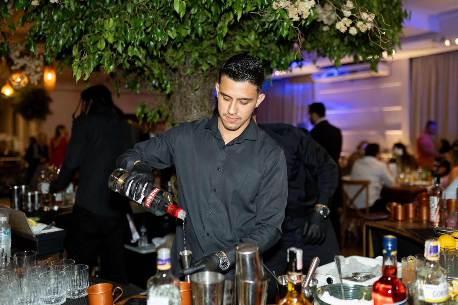 Bartender in black shirt pouring a drink at an elegant event bar with bottles and glassware on display.