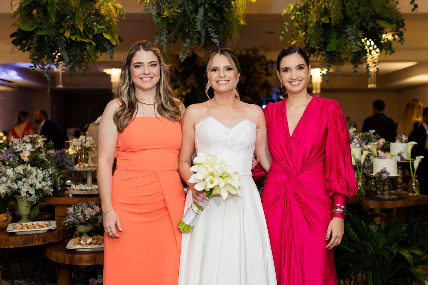 Bride in white gown poses with two bridesmaids in orange and pink dresses, holding a bouquet at a wedding reception.
