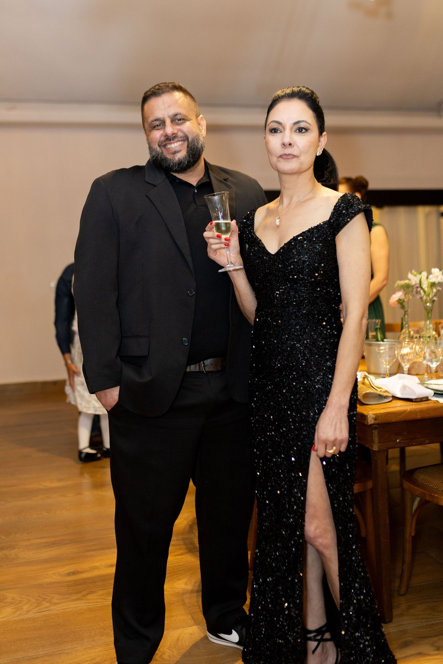 Elegant couple in black formal attire posing at a gala event, man holding champagne glass, woman in sequined gown.