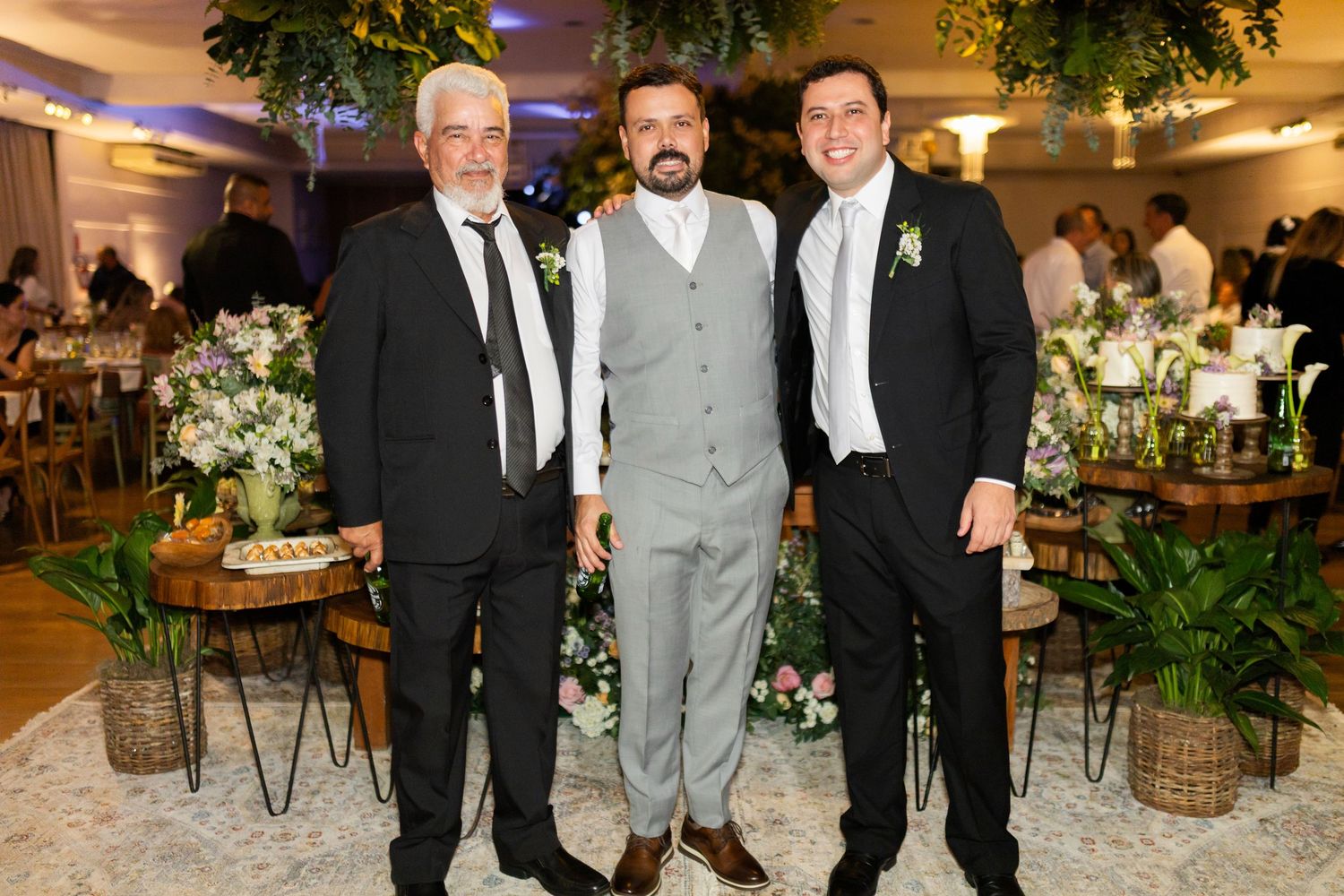 Three men in formal suits pose together at an elegant wedding reception with floral decorations.