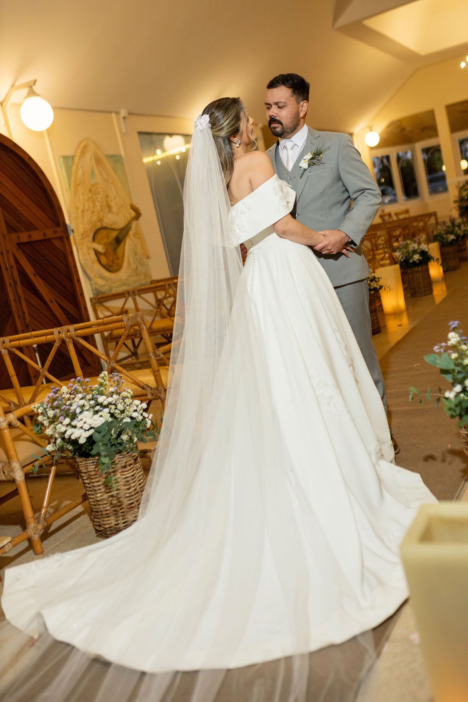 Bride in white gown with long veil and groom in gray suit share a romantic moment at an elegant wedding venue.