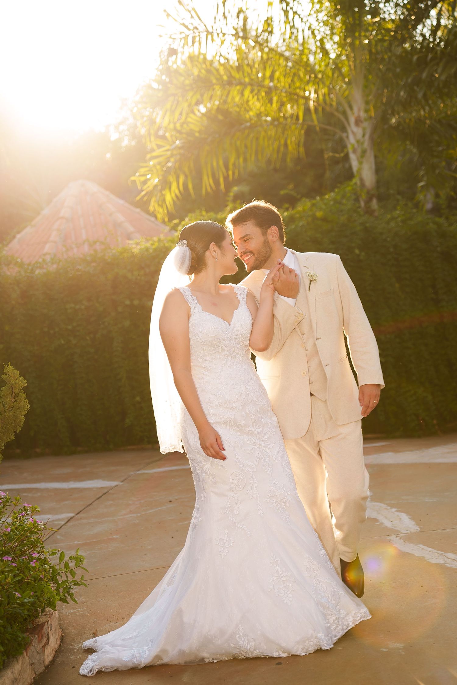 Bride in lace gown and groom in ivory suit share a romantic moment outdoors in golden sunset light.