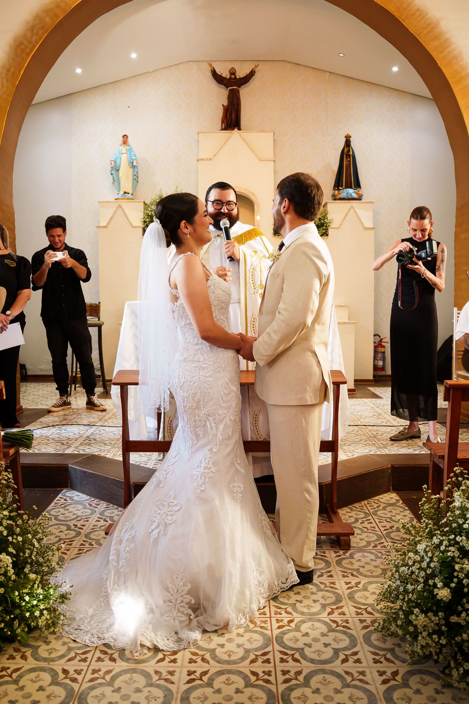 Bride and groom exchange vows at a Catholic church altar, surrounded by floral arrangements and wedding photographers.