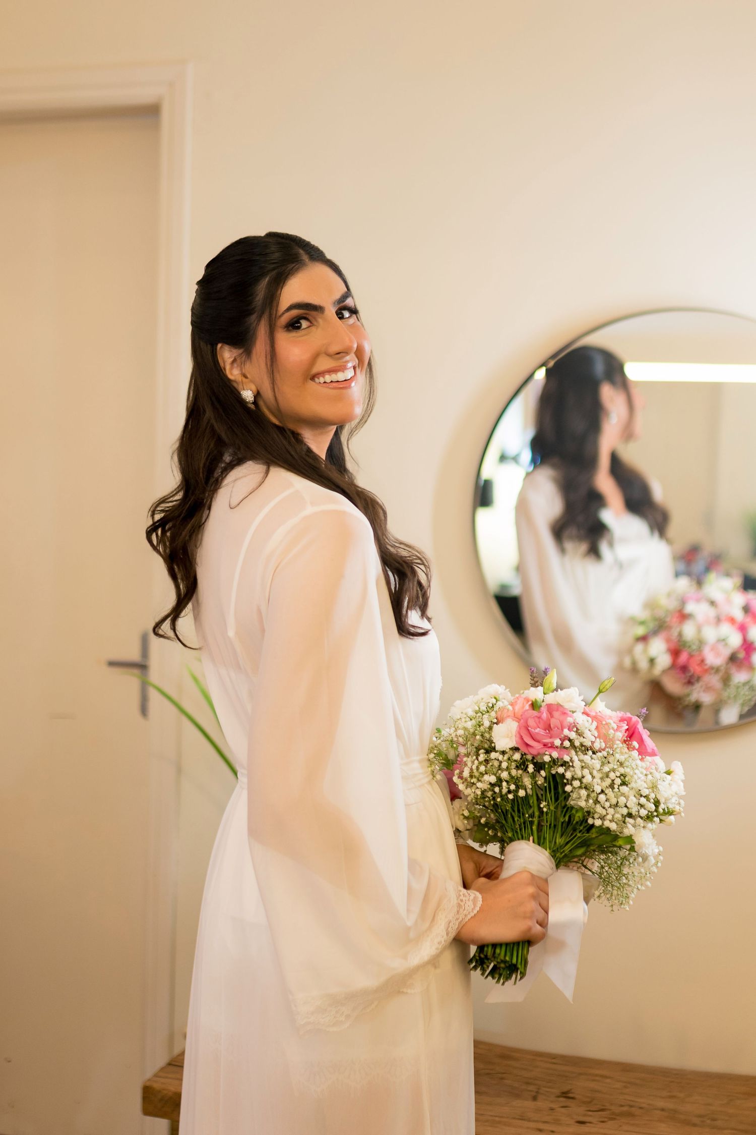 Smiling bride in white robe holding pink and white flower bouquet, with mirror reflection in background.