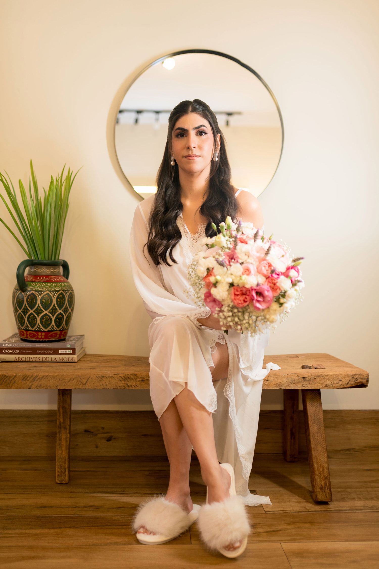 Bride in white robe and fluffy slippers holds floral bouquet, seated on wooden bench before round mirror.