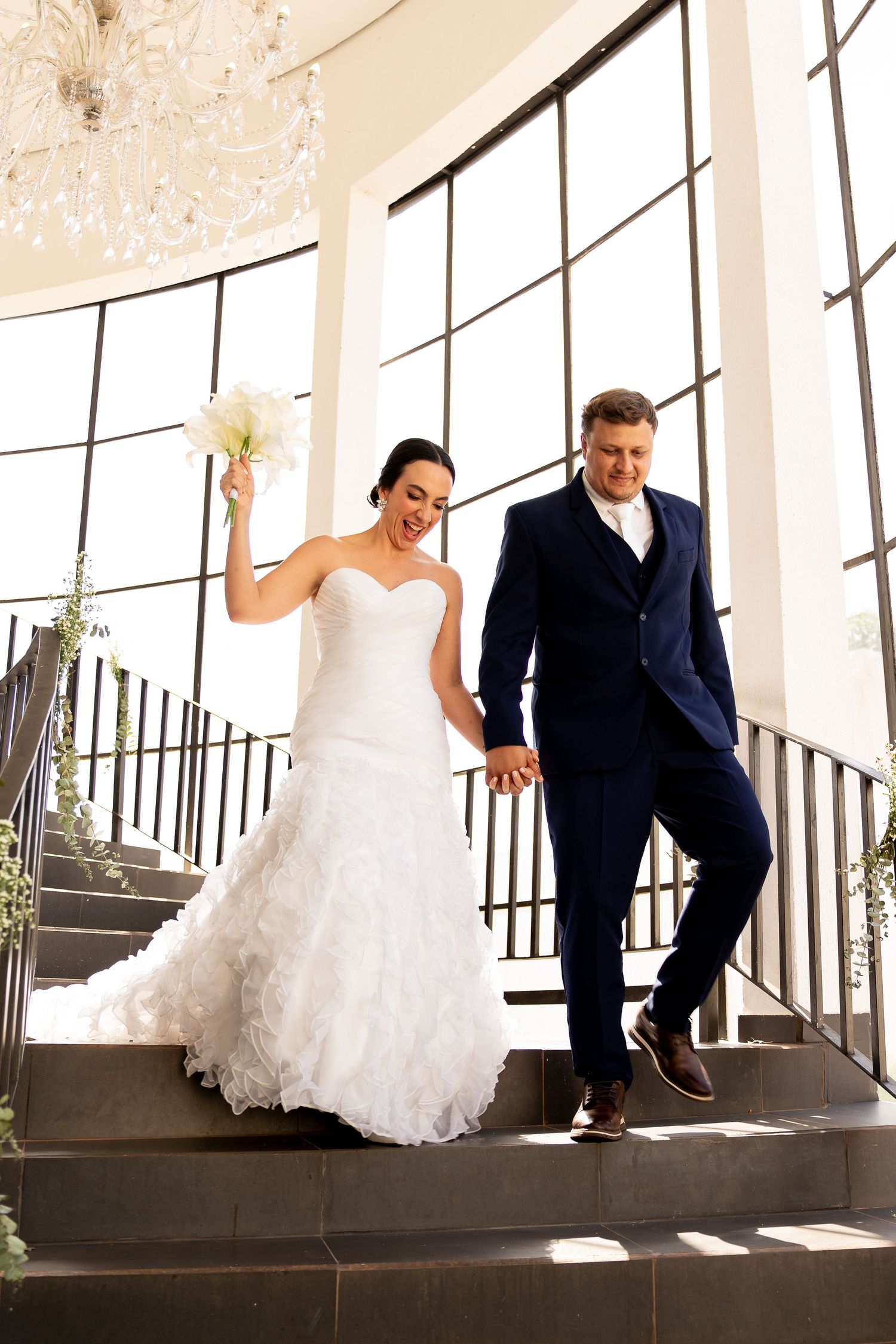 Bride and groom descend staircase, bride in white gown holding bouquet, groom in navy suit, elegant venue.
