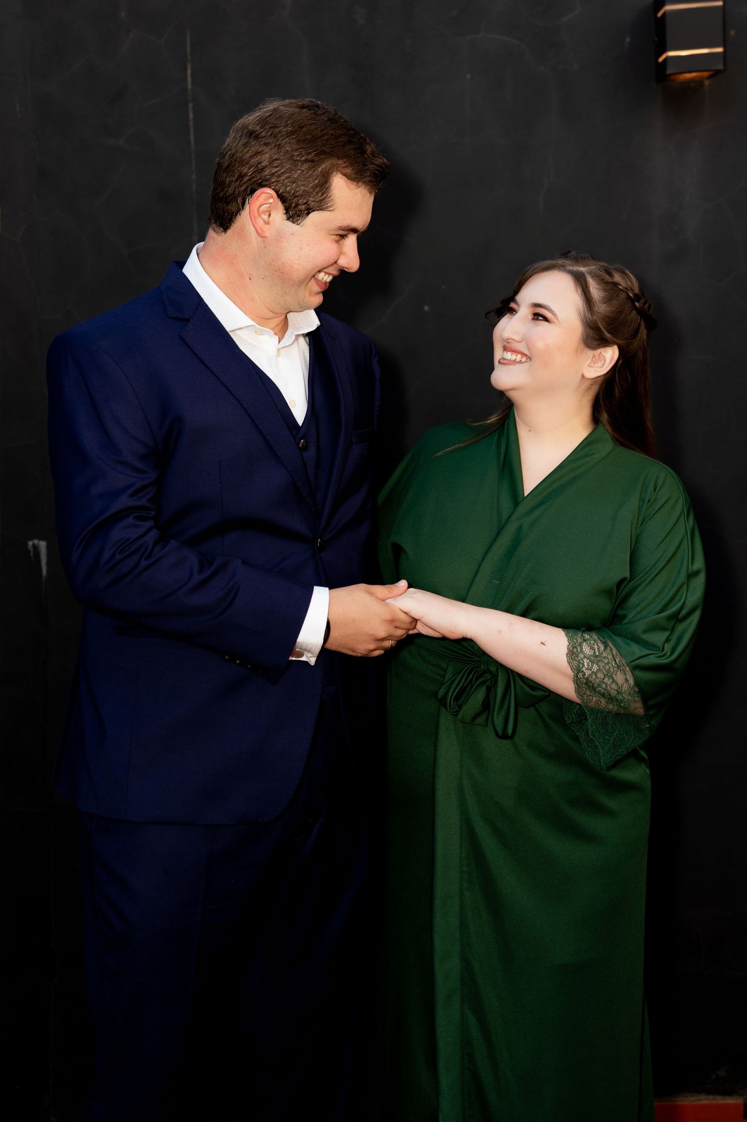 A smiling couple holding hands, man in navy suit and woman in green dress, posing against a dark wall.