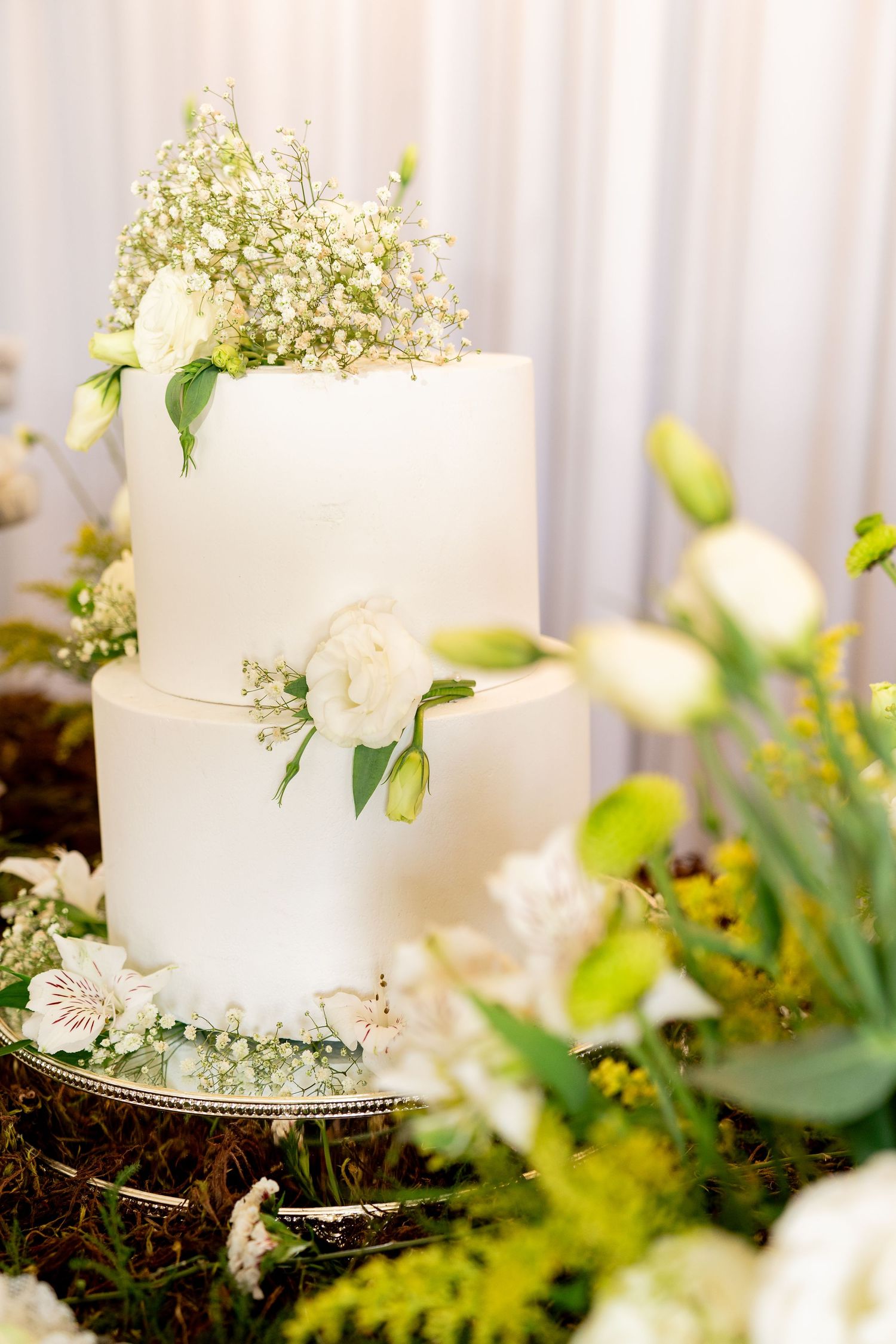 Elegant two-tier white wedding cake adorned with fresh white flowers and greenery, surrounded by floral arrangements.