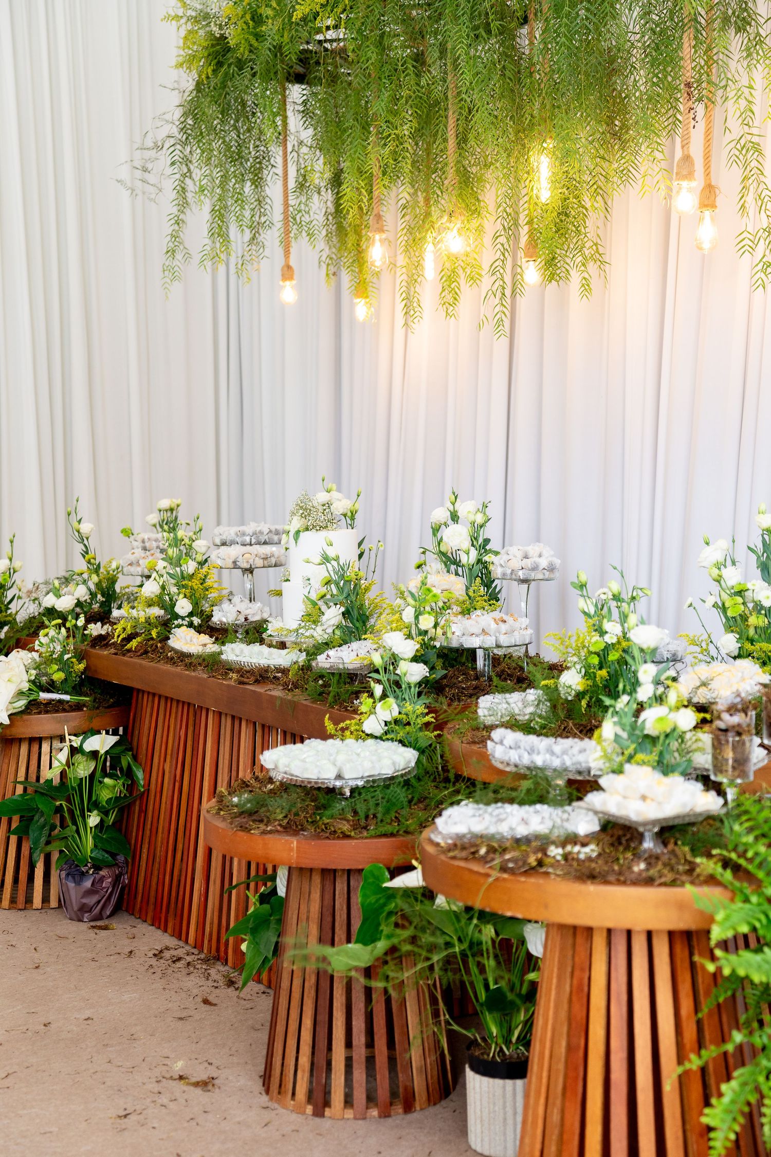 Elegant wedding dessert table with white flowers, greenery, wooden stands, and warm Edison bulb lighting.