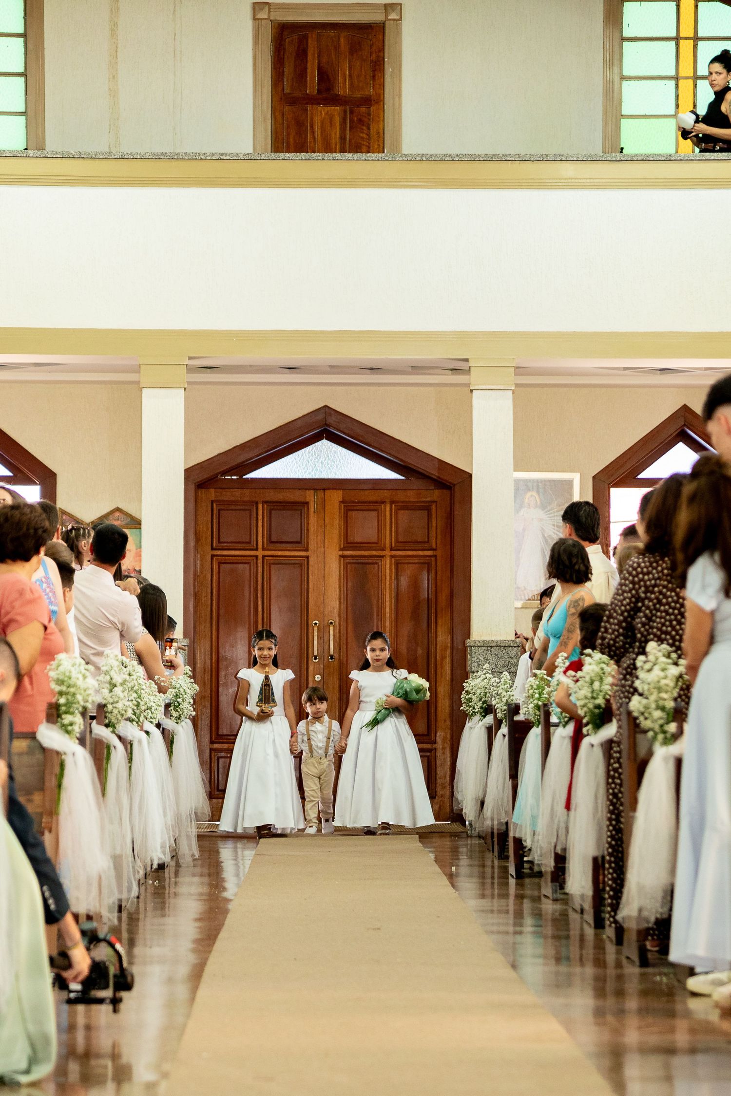 Flower girls in white dresses walk down church aisle at wedding ceremony.