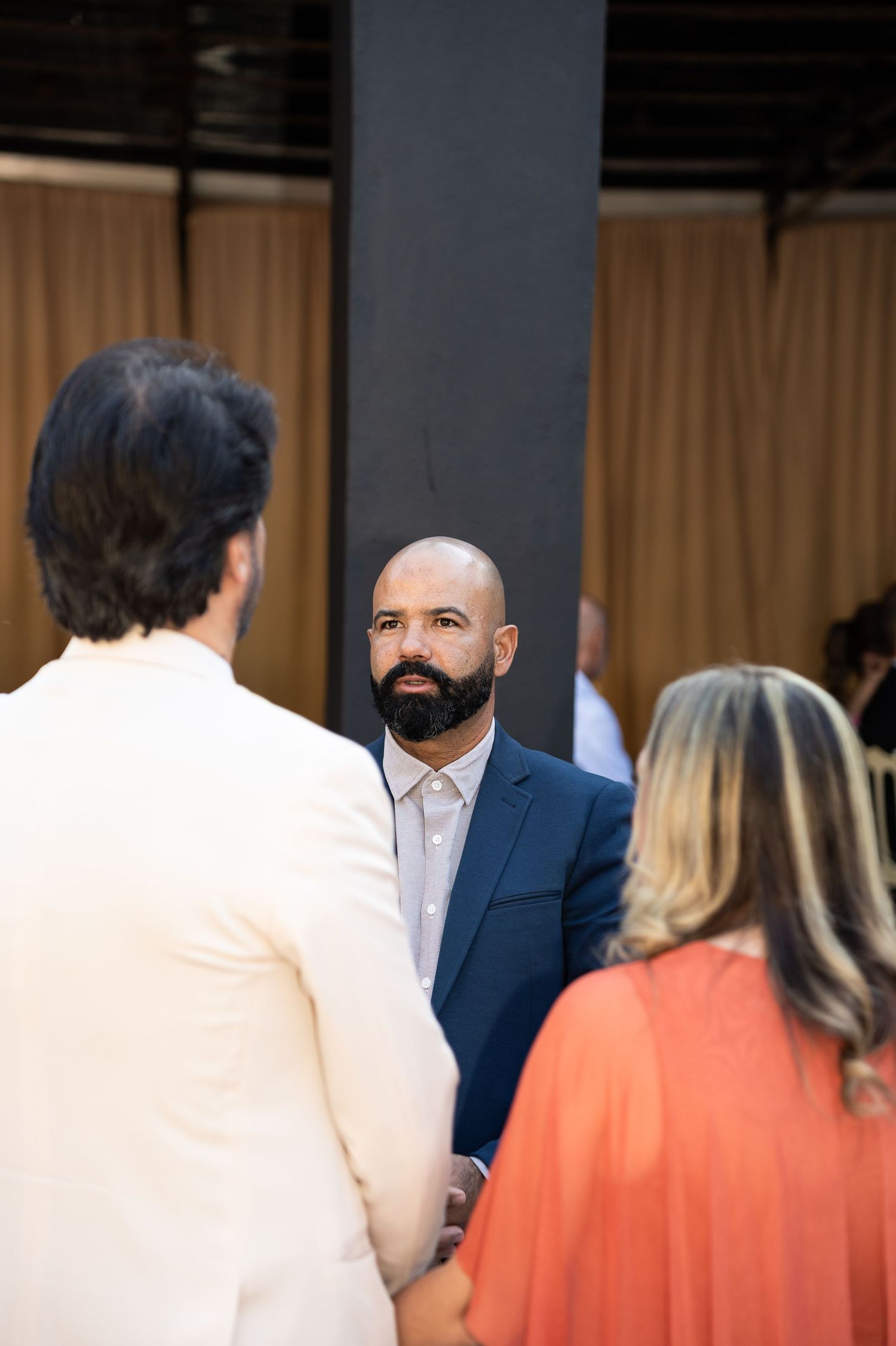 Bald man with beard in navy suit listening attentively during an indoor event with curtain backdrop.