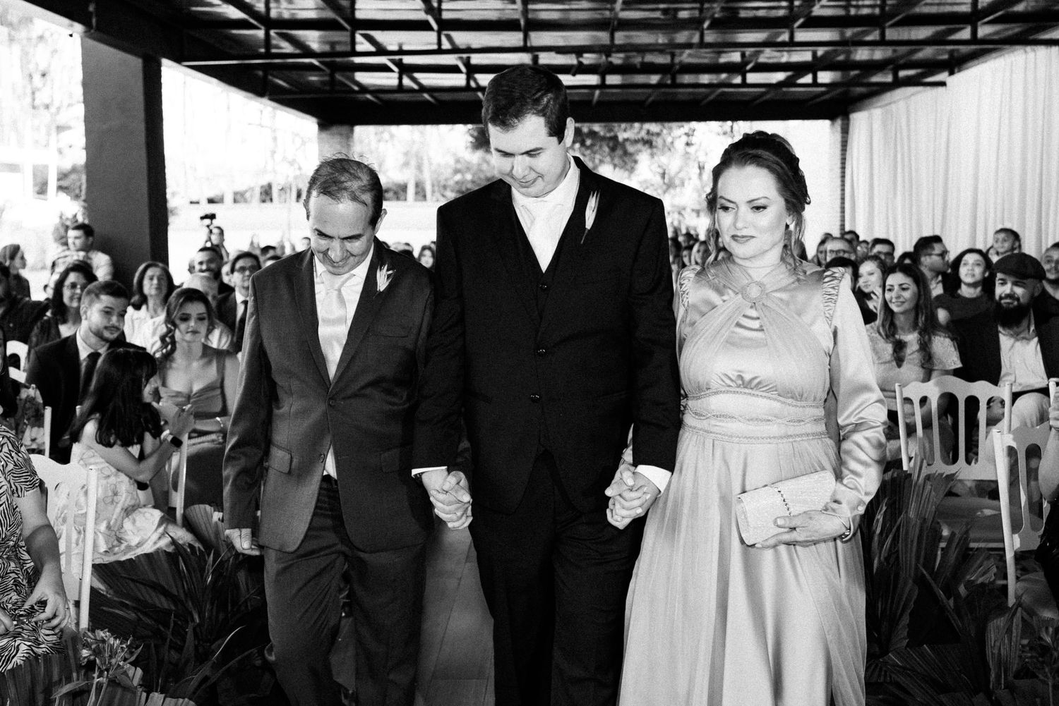 Black and white wedding photo of groom walking down aisle with two family members holding hands at outdoor ceremony.