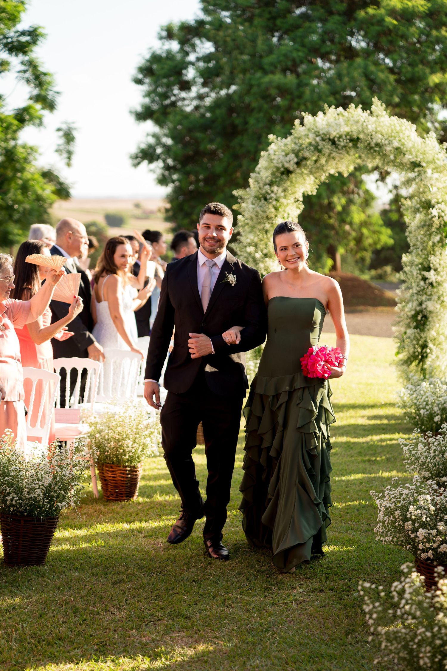 Couple walks down outdoor wedding aisle under floral arch, woman in green gown holding pink bouquet, guests applauding.