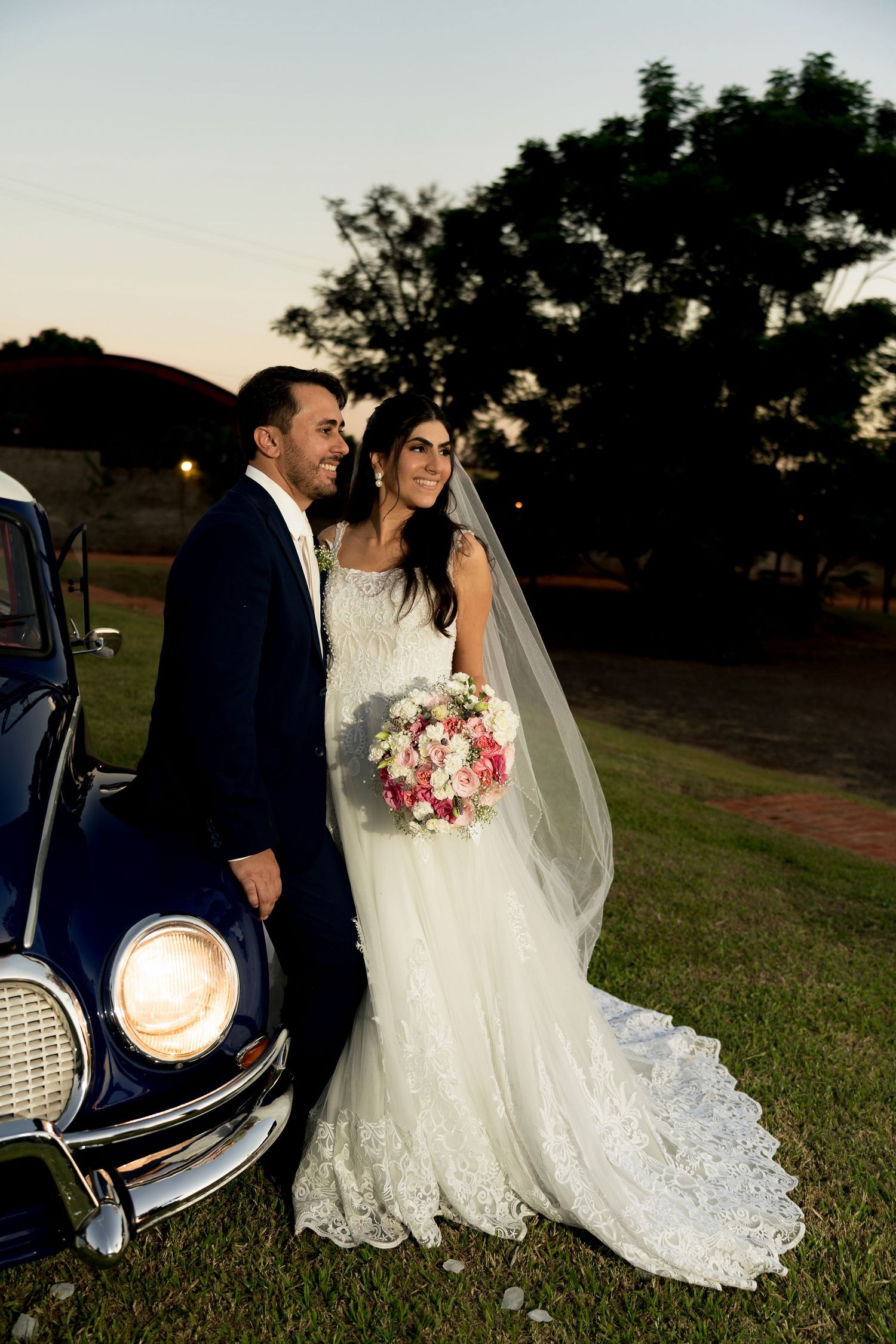 Bride and groom pose beside a vintage car at dusk, bride holding pink bouquet in lace wedding gown with veil.