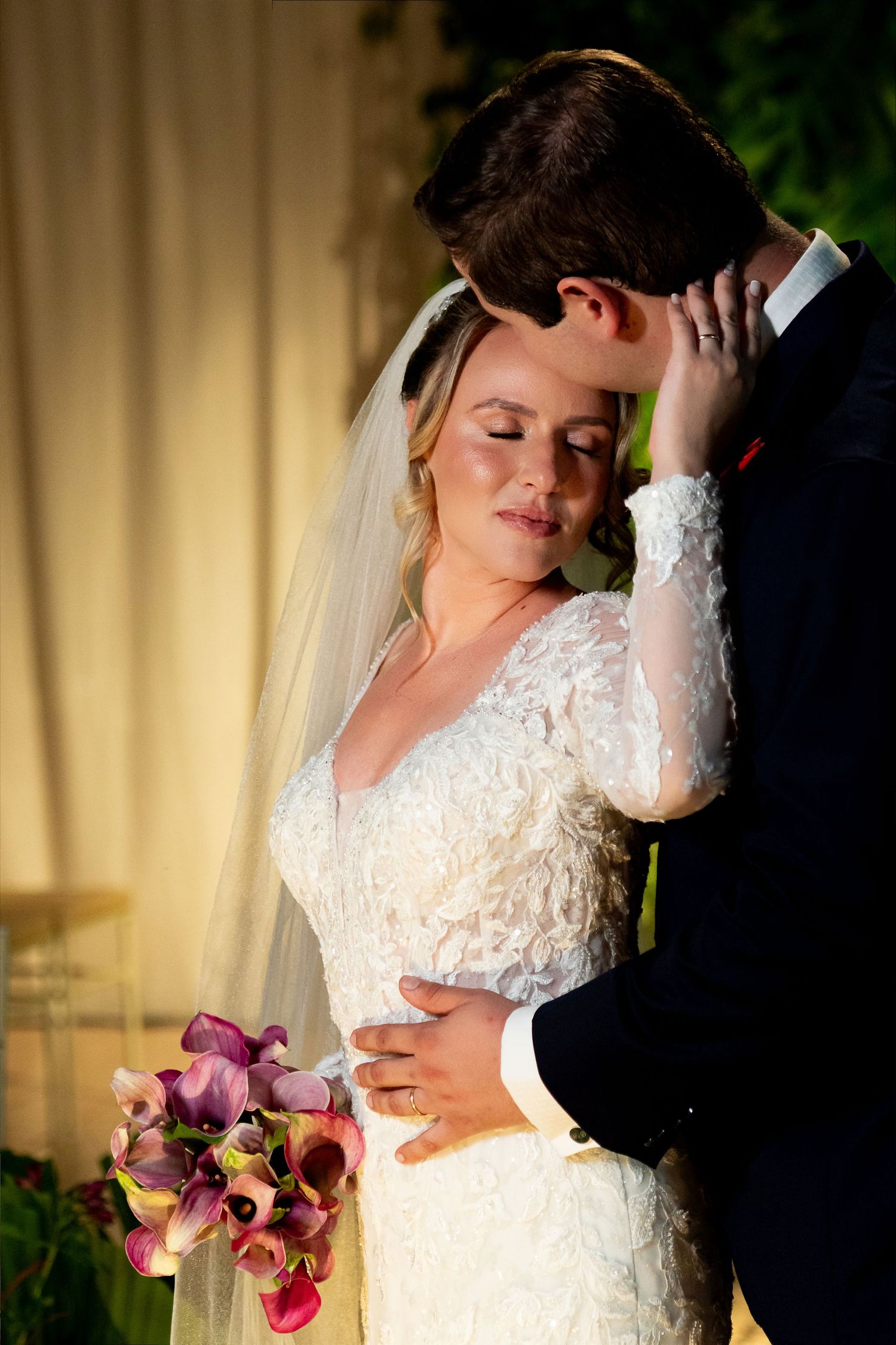 Bride in lace gown holding purple calla lilies embraces groom in navy suit during romantic wedding ceremony.