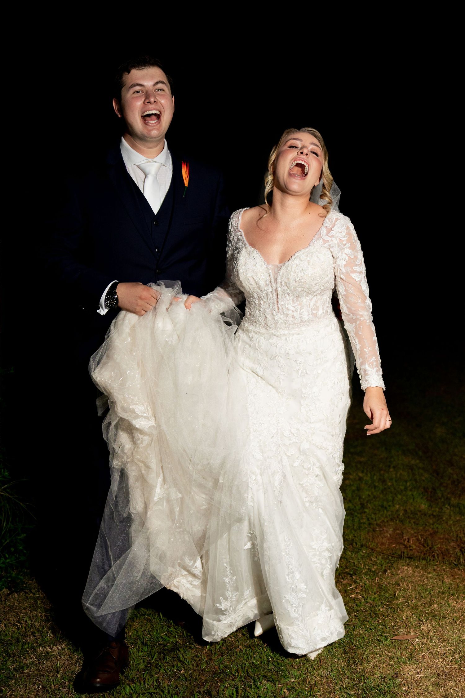 Joyful bride in lace gown and groom in navy suit laughing together outdoors at night during wedding celebration.