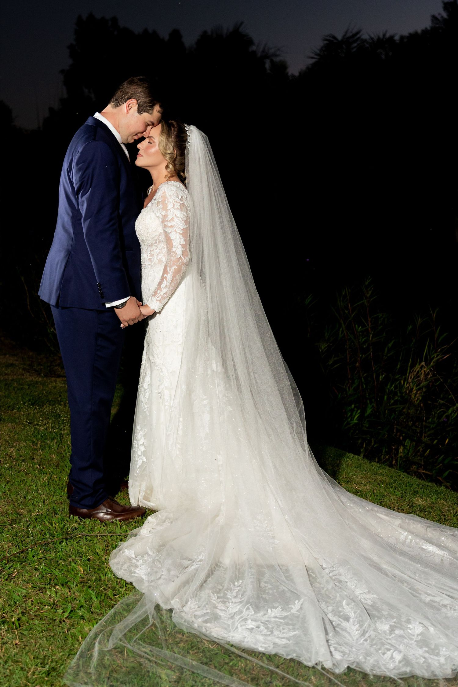 Bride and groom share a romantic moment at night, her lace gown and veil glowing against the dark backdrop.