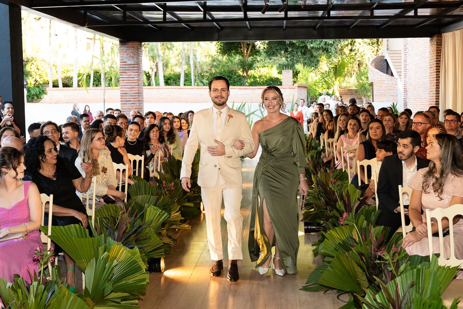 Elegant outdoor wedding ceremony with groom in white suit and bridesmaid in green dress walking down aisle.