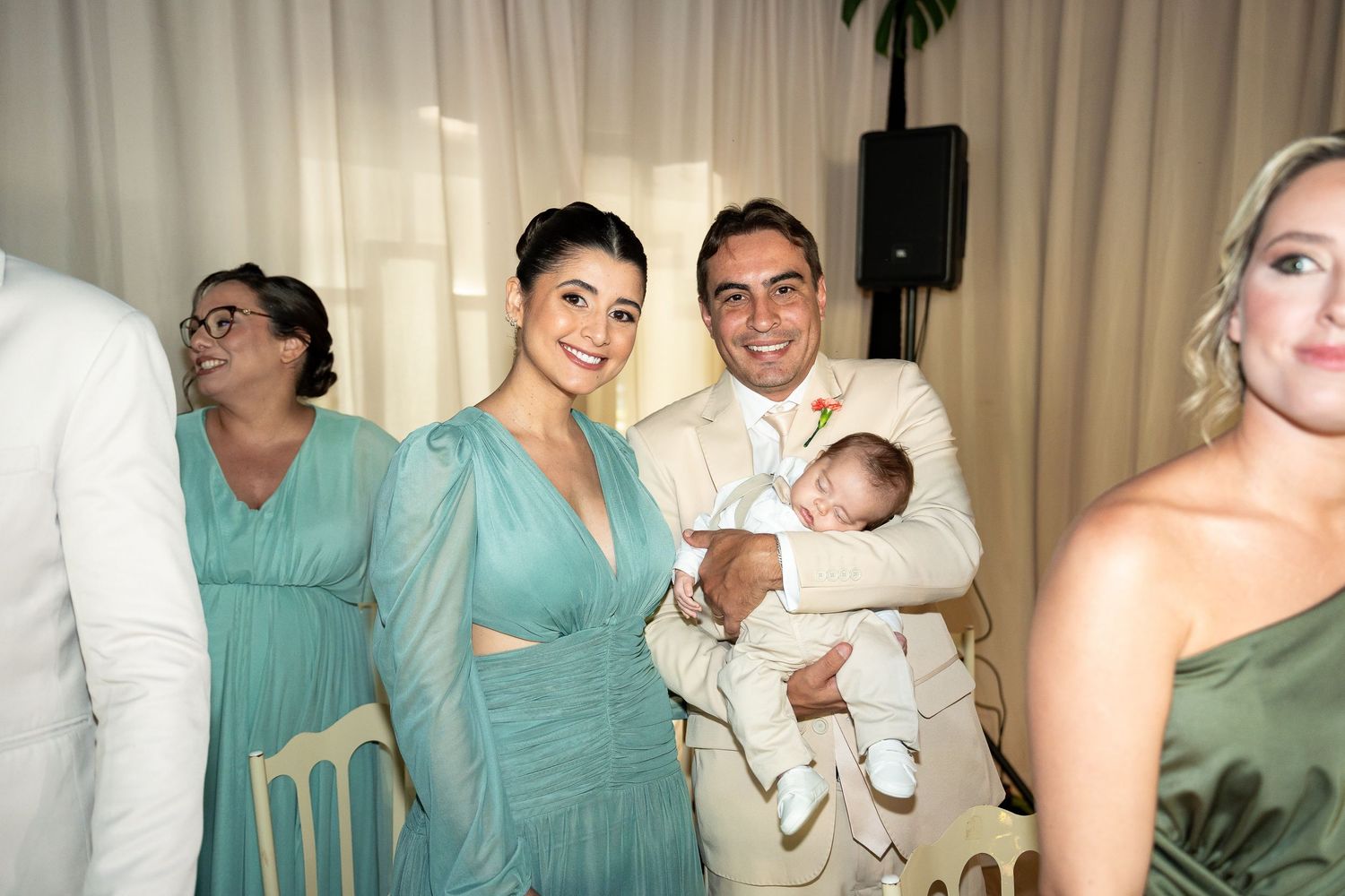 Couple in elegant attire holding a baby at a wedding reception with guests in teal dresses in background.